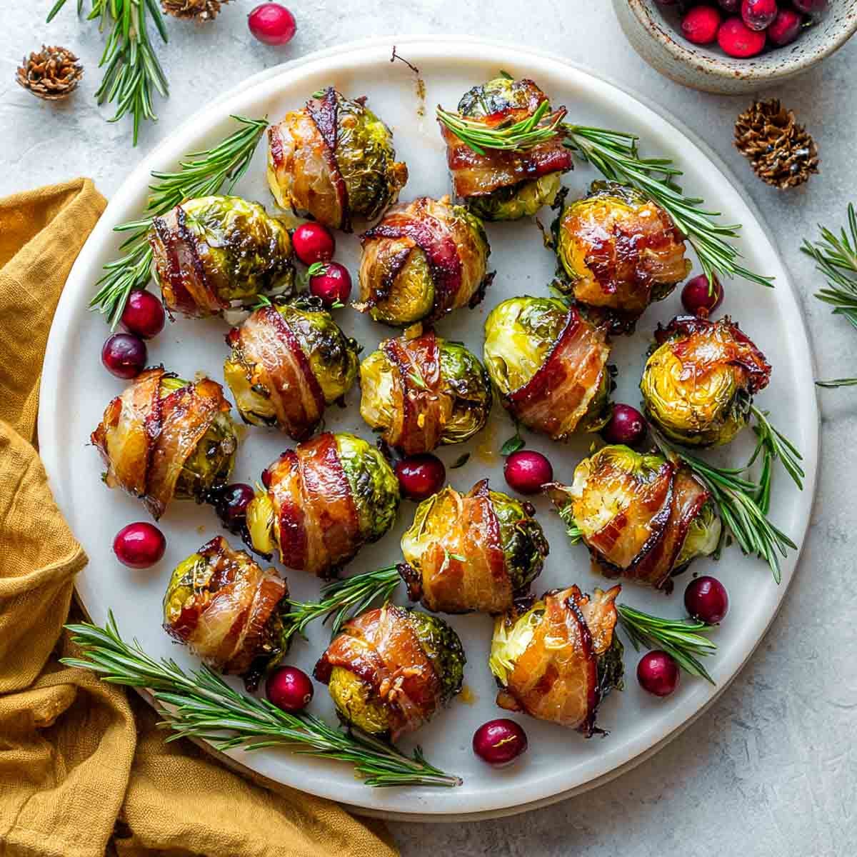 Brussels sprouts wrapped in crispy bacon with maple glaze, arranged on a white platter with rosemary and cranberries.