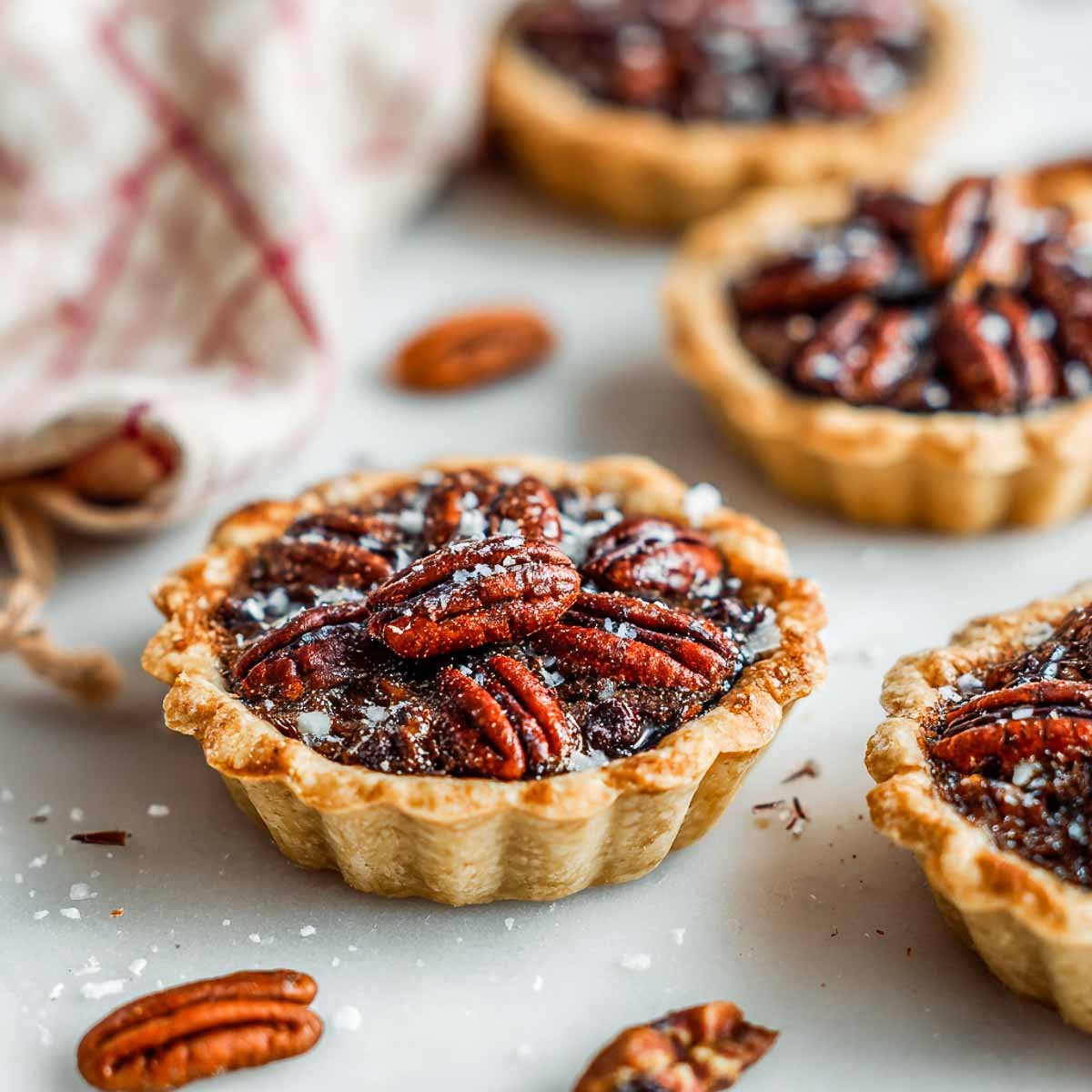 Mini gluten-free chocolate pecan pies with golden crusts, glossy pecans, and a sprinkle of sea salt on top.