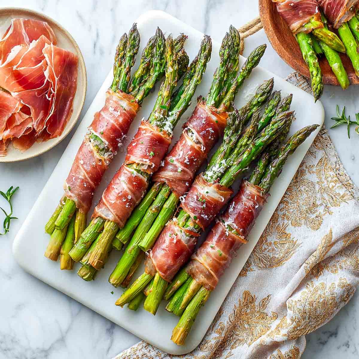 Asparagus bundles wrapped in crispy roasted prosciutto on a platter with raw prosciutto slices on the side.