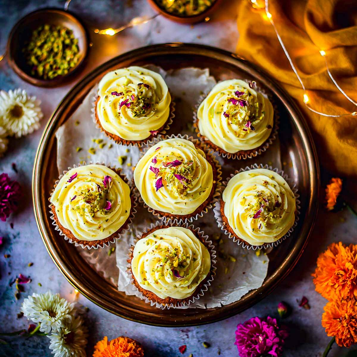 Tray of Rasmalai cupcakes decorated with saffron frosting, pistachios, rose petals.