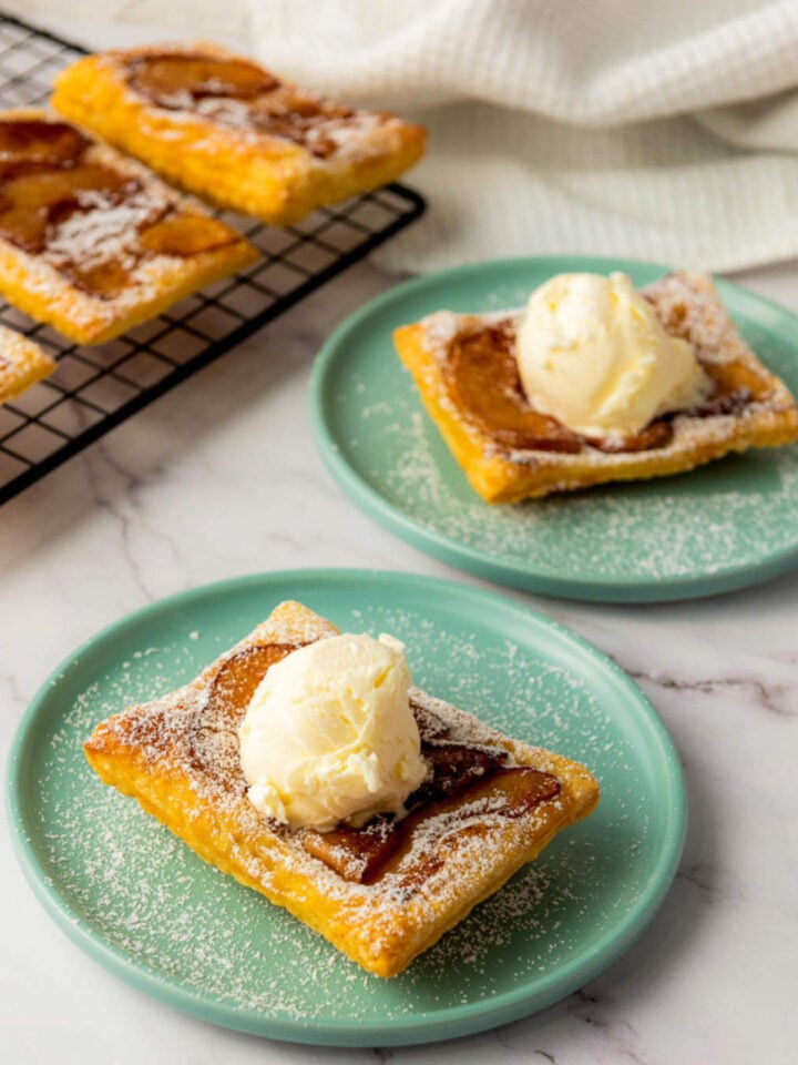 Two upside down apple tarts with ice cream on teal plates beside a cooling rack with extra tarts in the background.