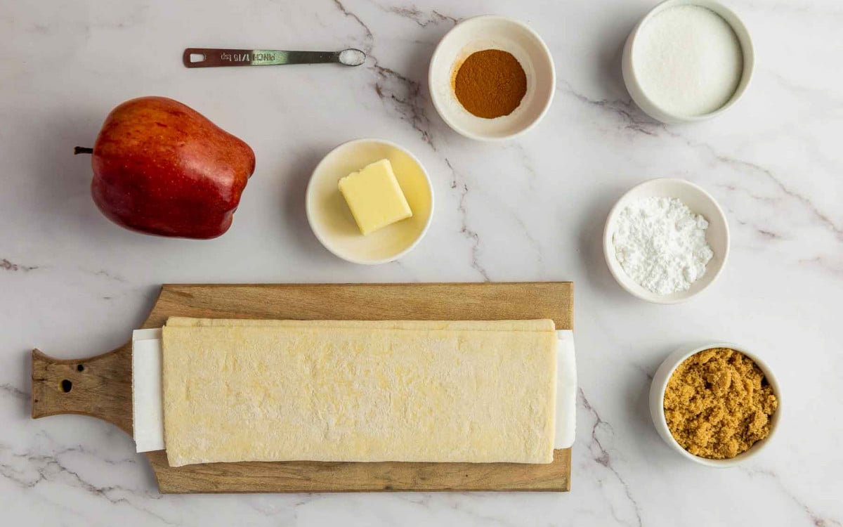 Ingredients for upside down apple tart including puff pastry, apple, butter, sugars, and cinnamon on a marble surface.