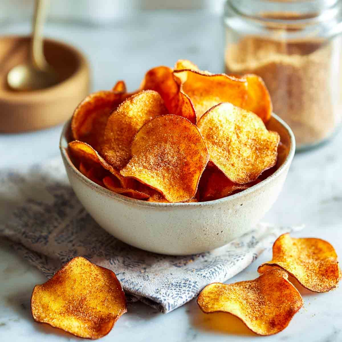 A bowl of golden sweet potato chips dusted with cinnamon sugar, on a muted patterned napkin with a cinnamon jar in the background.