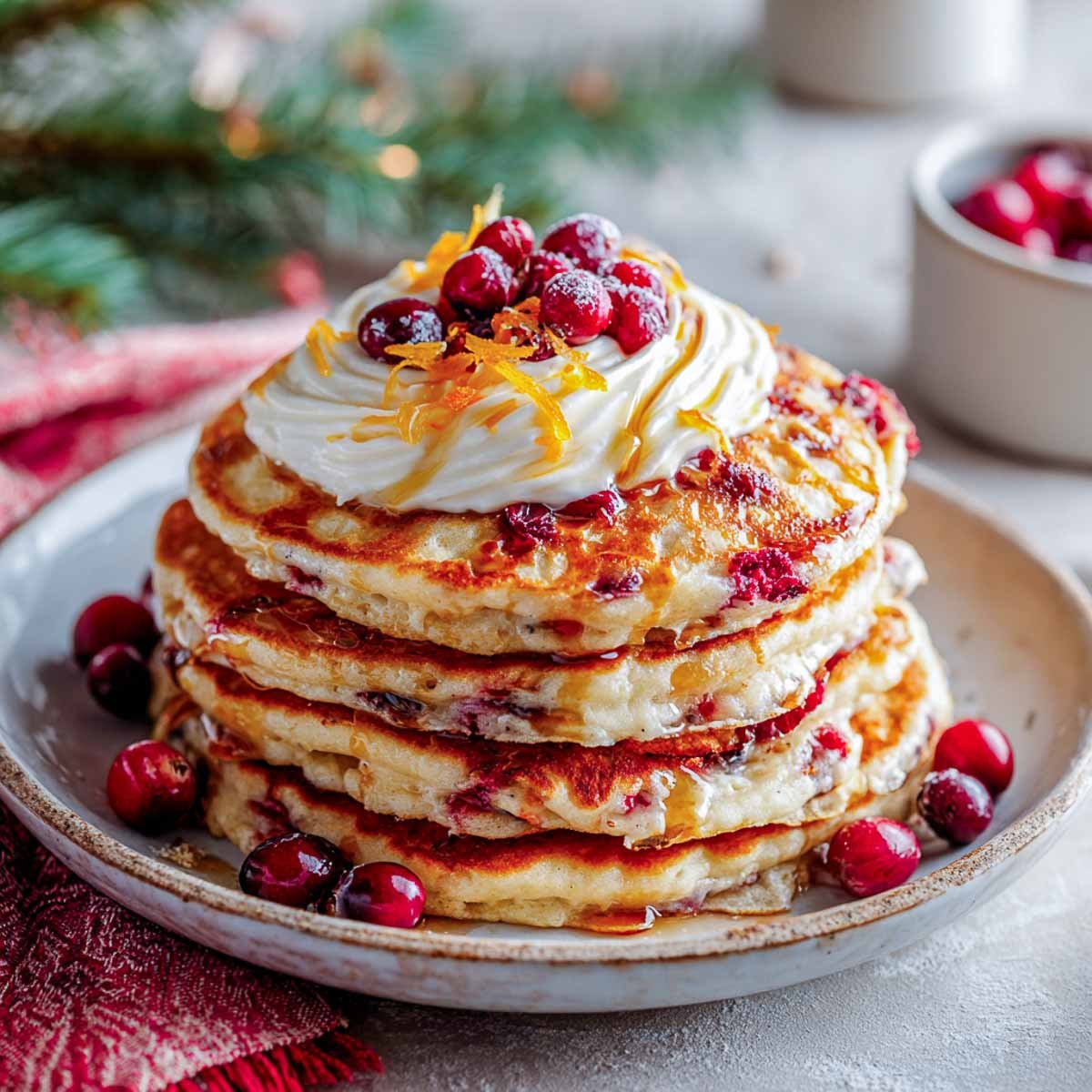 Stack of cranberry oat pancakes topped with orange zest yogurt and cranberries on a Christmas breakfast table.