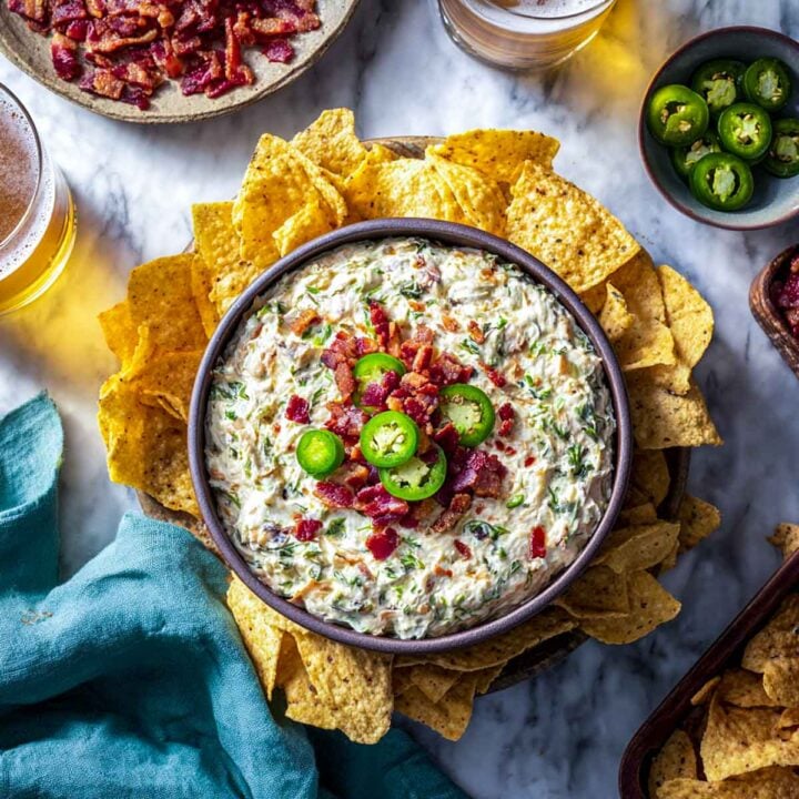 Bowl of creamy jalapeño popper dip surrounded by tortilla chips, topped with bacon and jalapeño slices on a marble surface.