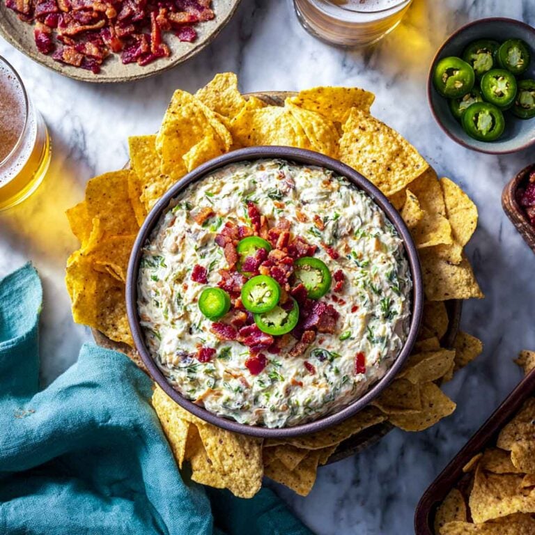 Bowl of creamy jalapeño popper dip surrounded by tortilla chips, topped with bacon and jalapeño slices on a marble surface.