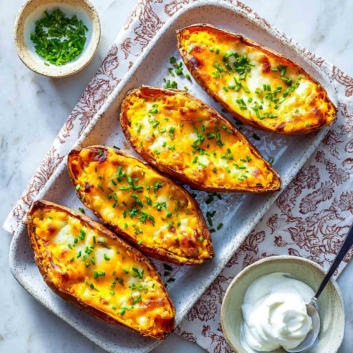 Four air-fried sweet potato skins topped with melted cheese and chives on a rectangular serving plate with sour cream on the side.