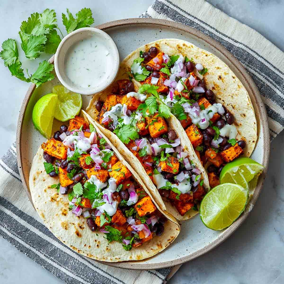 Top-down view of two soft tacos filled with sweet potatoes, black beans, red onion, cilantro, and lime crema on a ceramic plate with sliced limes nearby.