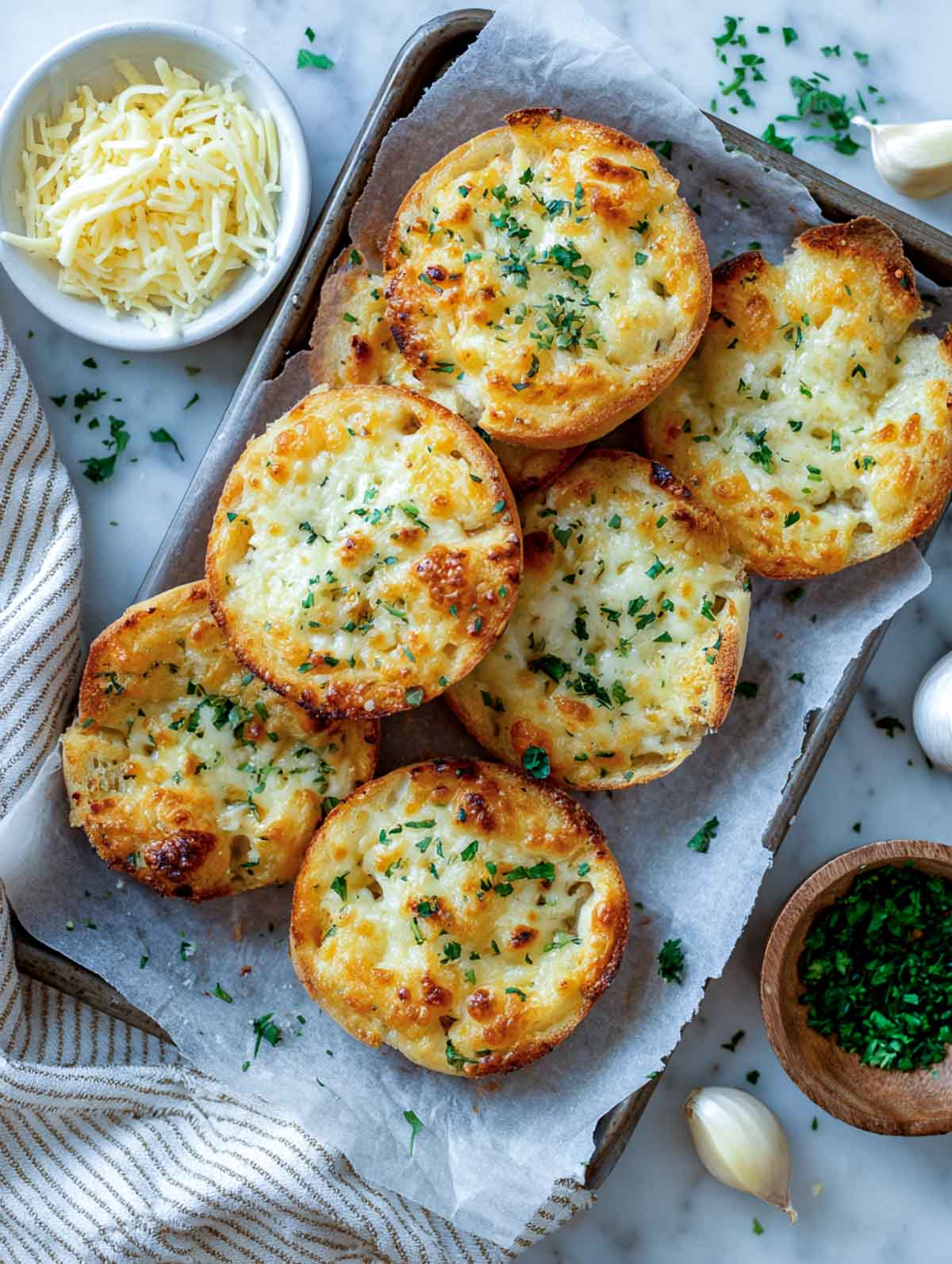 Golden cheddar garlic bread rounds on a parchment-lined tray with shredded cheese, herbs, and garlic on a marble counter.