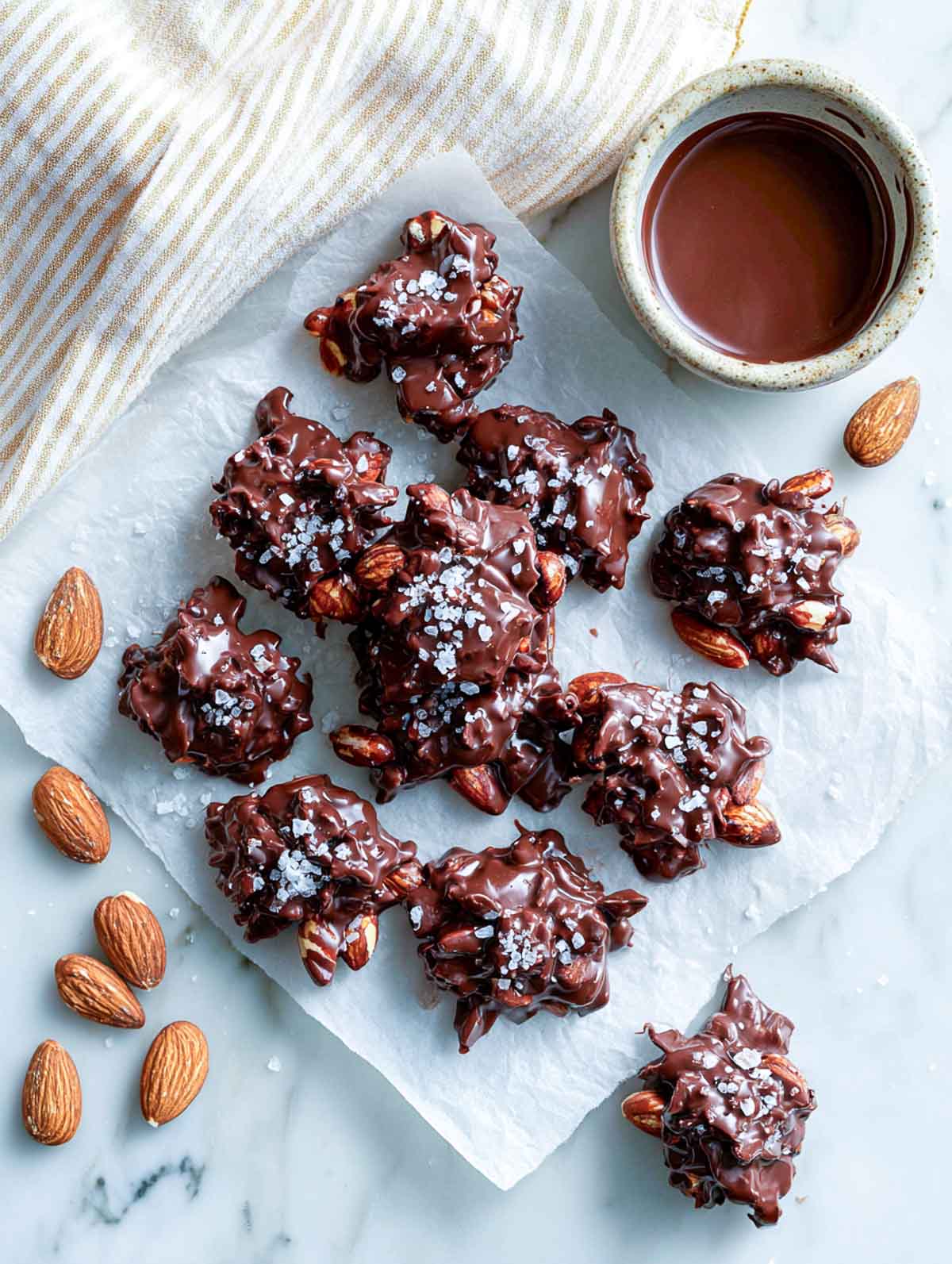 Chocolate almond clusters topped with sea salt on parchment paper beside a cup of melted chocolate.