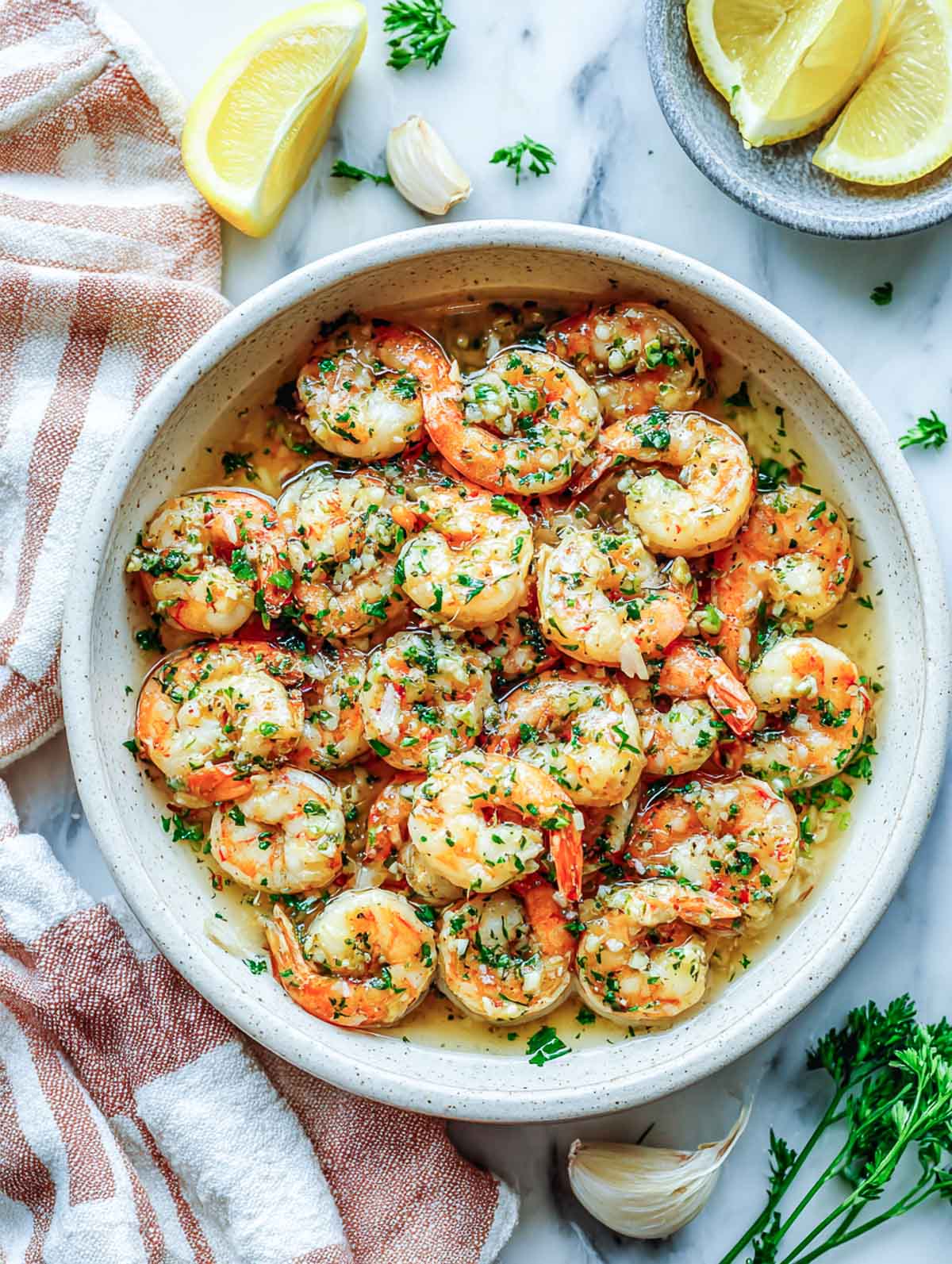 Garlic butter shrimp in a cream-colored bowl with lemon wedges and parsley on a marble countertop.