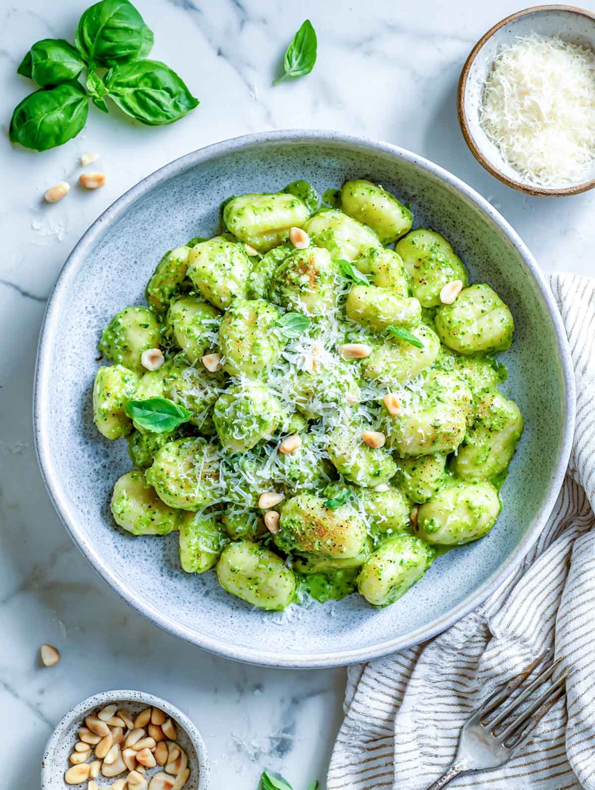 Creamy pesto gnocchi in a gray-blue bowl topped with parmesan, pine nuts, and basil on a marble counter.