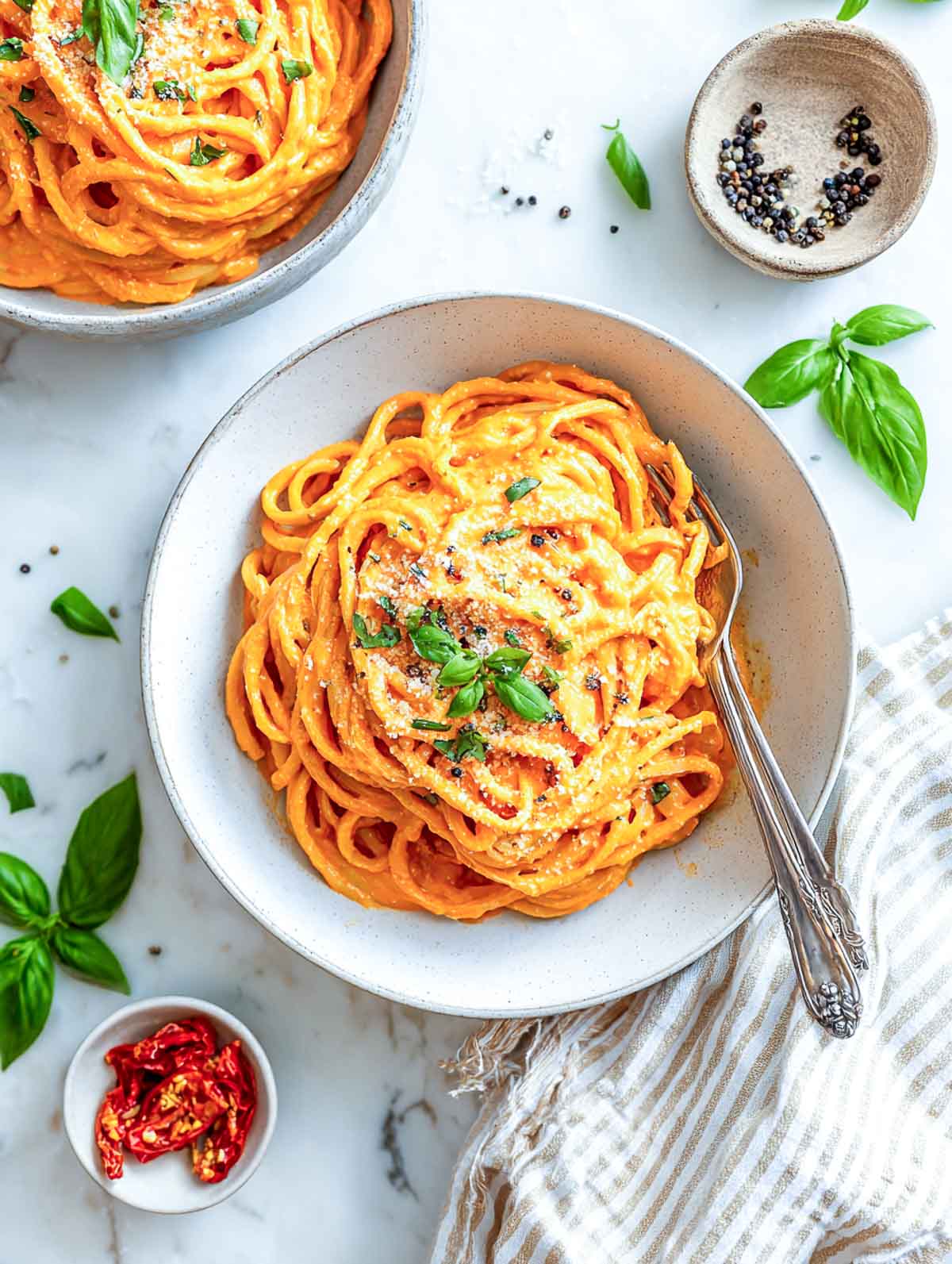 Creamy roasted red pepper pasta in an off-white bowl topped with basil and parmesan on a marble counter.