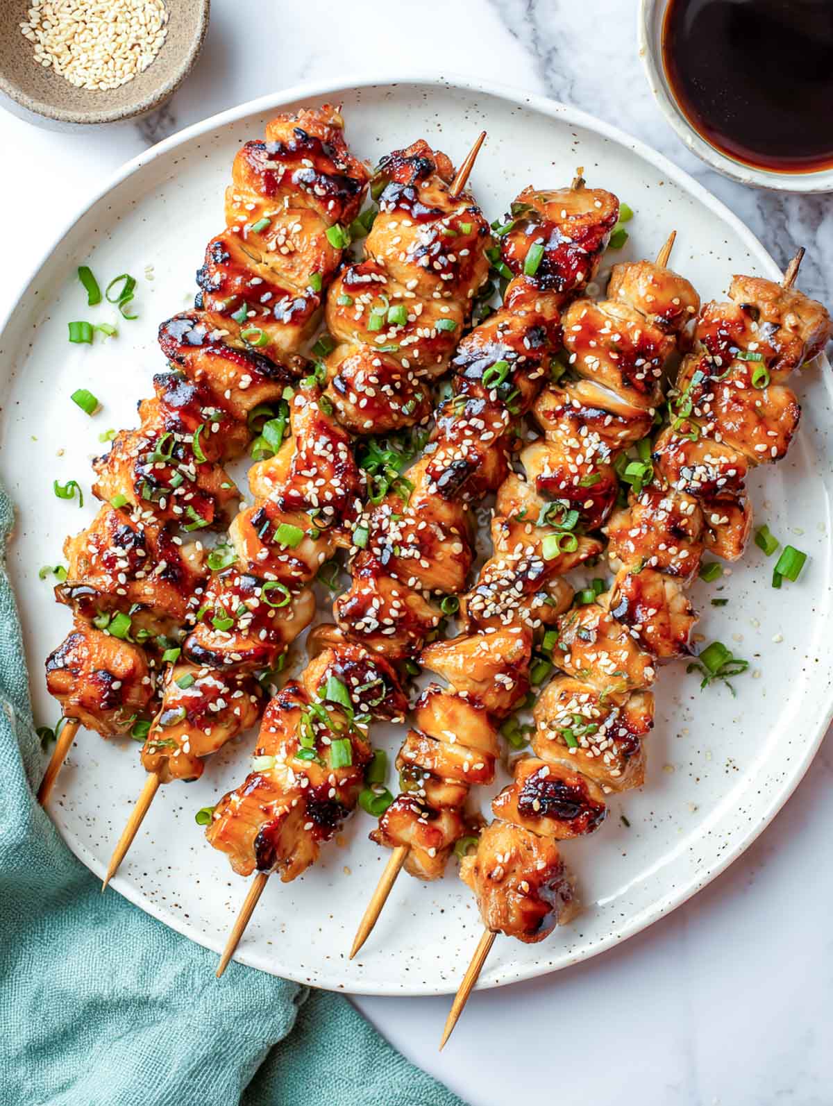 Teriyaki chicken skewers on a light gray plate, brushed with glaze and sprinkled with sesame seeds and scallions.