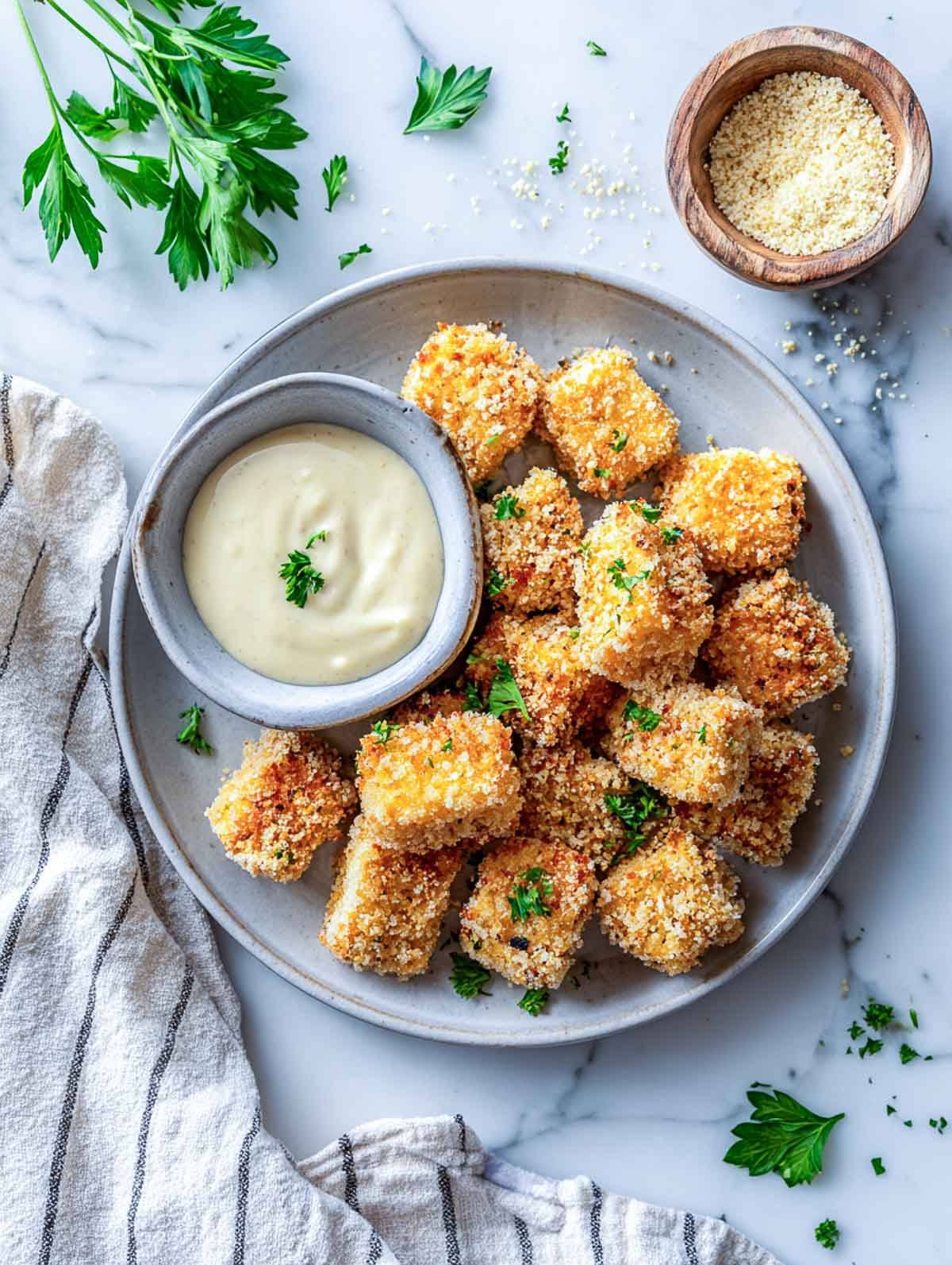 Golden crispy tofu nuggets on a gray plate with dipping sauce and parsley on a marble counter.