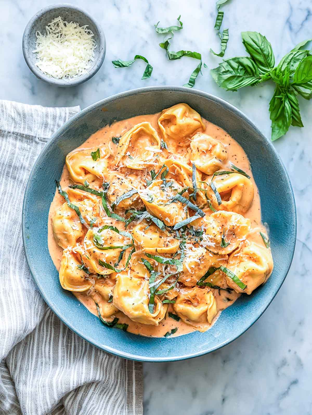 Creamy tomato tortellini in a muted blue bowl topped with basil ribbons and grated parmesan on a marble counter.
