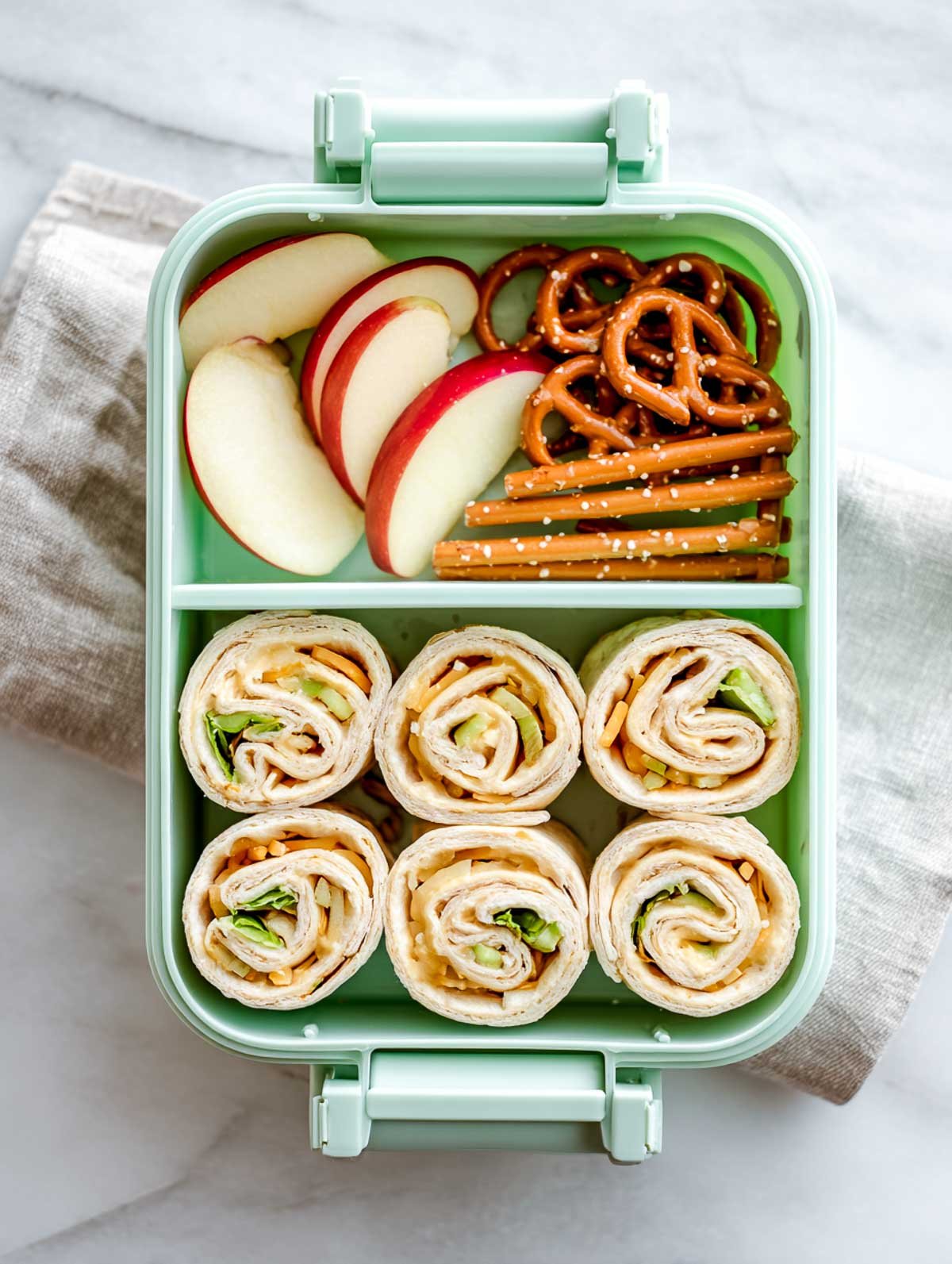 Apple cheddar roll-ups in a mint-green lunchbox with apple slices and pretzels on a marble surface.
