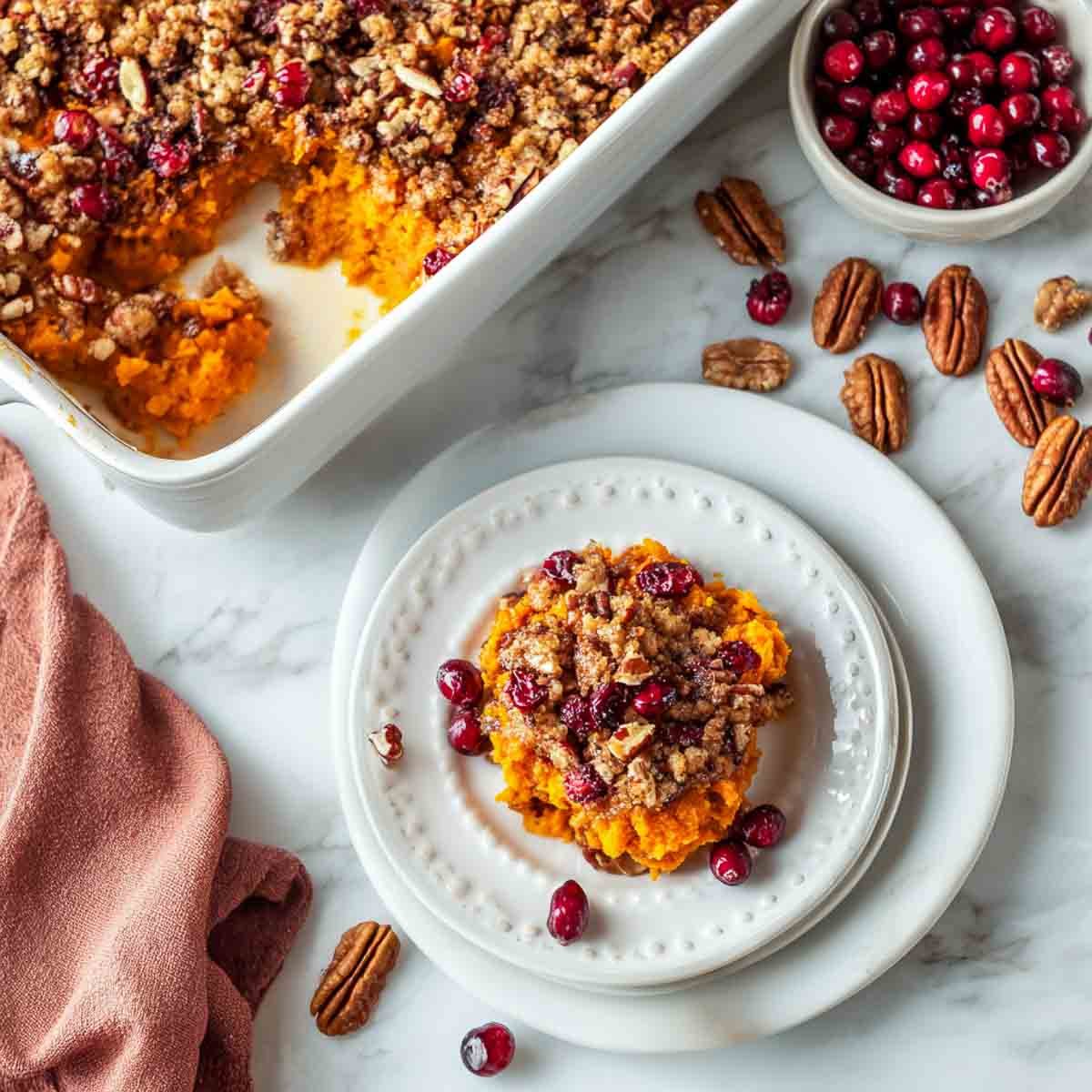 A plated serving of baked sweet potato casserole with cranberry pecan streusel, baking dish in the background.