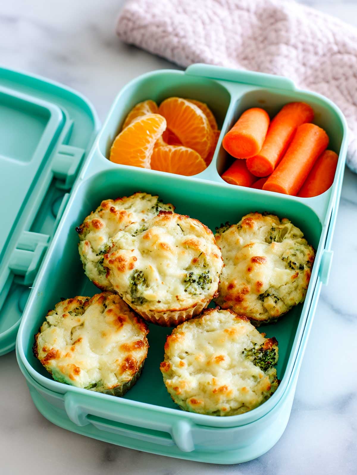 Cheesy broccoli bites packed in a mint-green lunchbox with mandarin orange segments and baby carrots on a marble surface.
