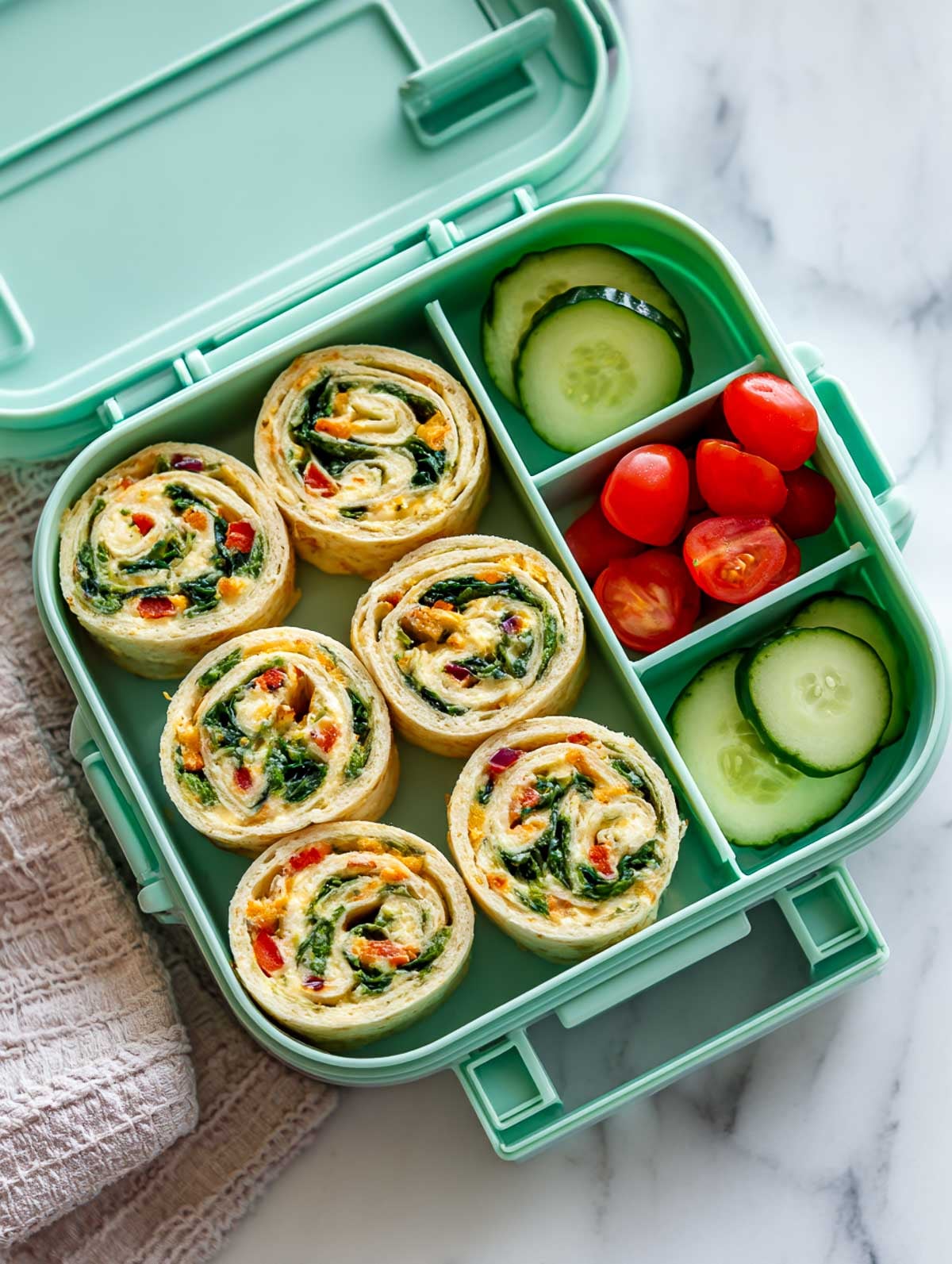 Cheese and veggie pinwheel roll-ups in a mint-green lunchbox with cucumber and cherry tomato sides on a marble surface.
