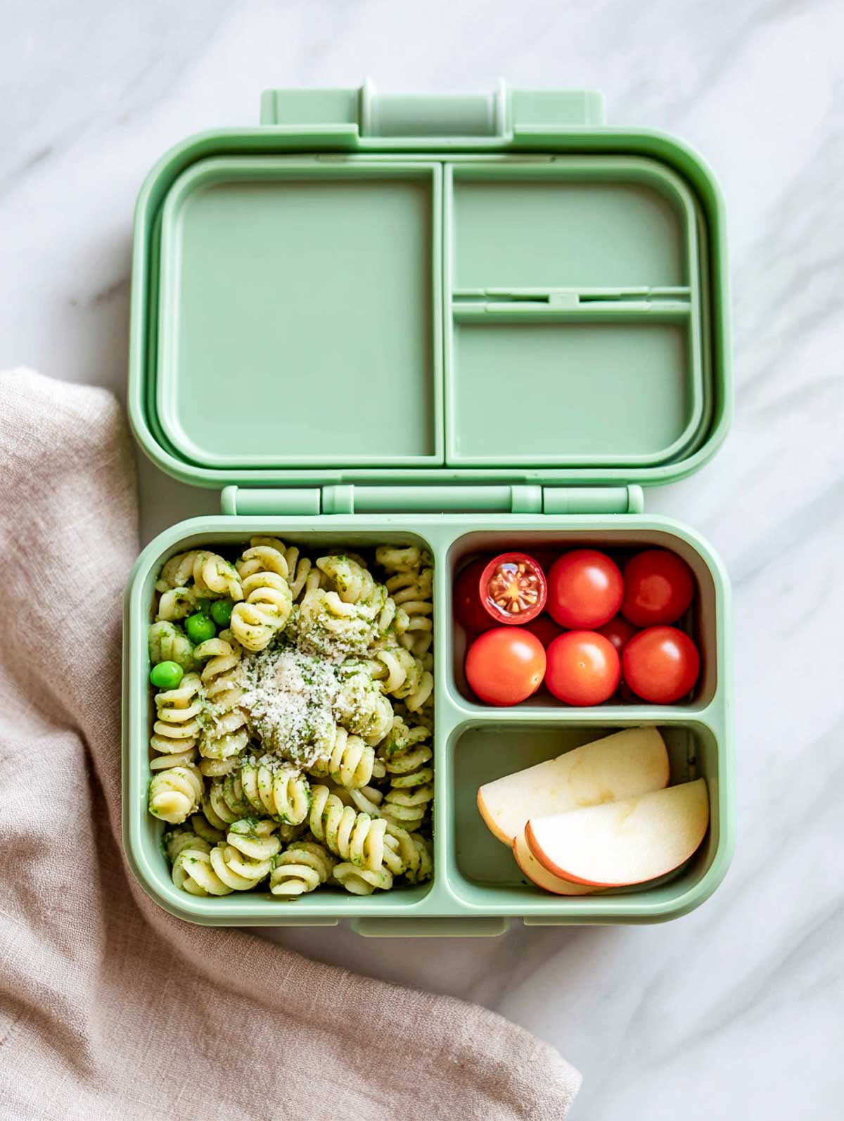 Cold pesto pasta twirls with peas and parmesan in a mint-green bento lunchbox with cherry tomatoes and apple slices.