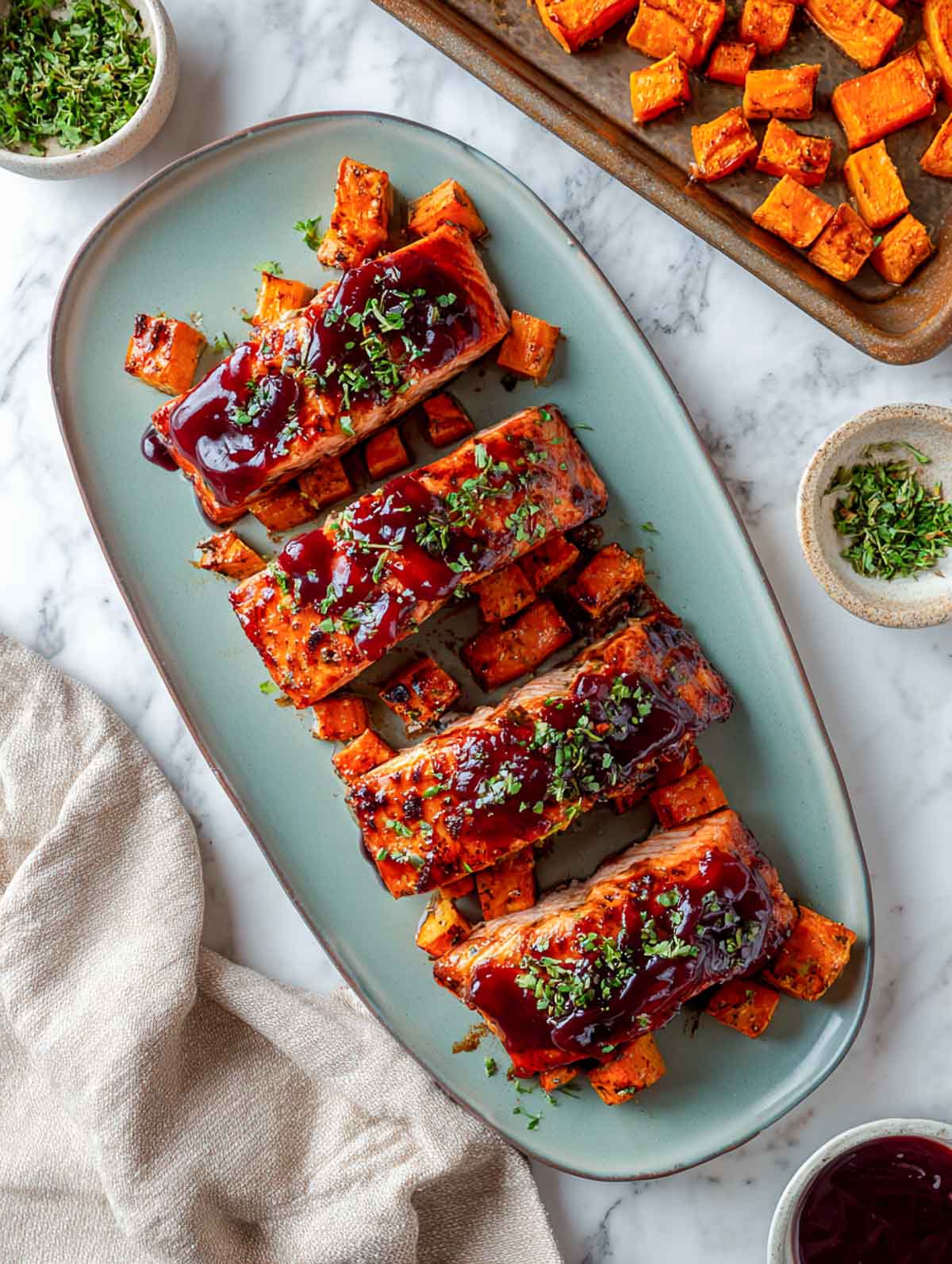 Top-down view of four cranberry-glazed salmon fillets arranged on a rectangular blue plate with roasted sweet potato cubes scattered around.