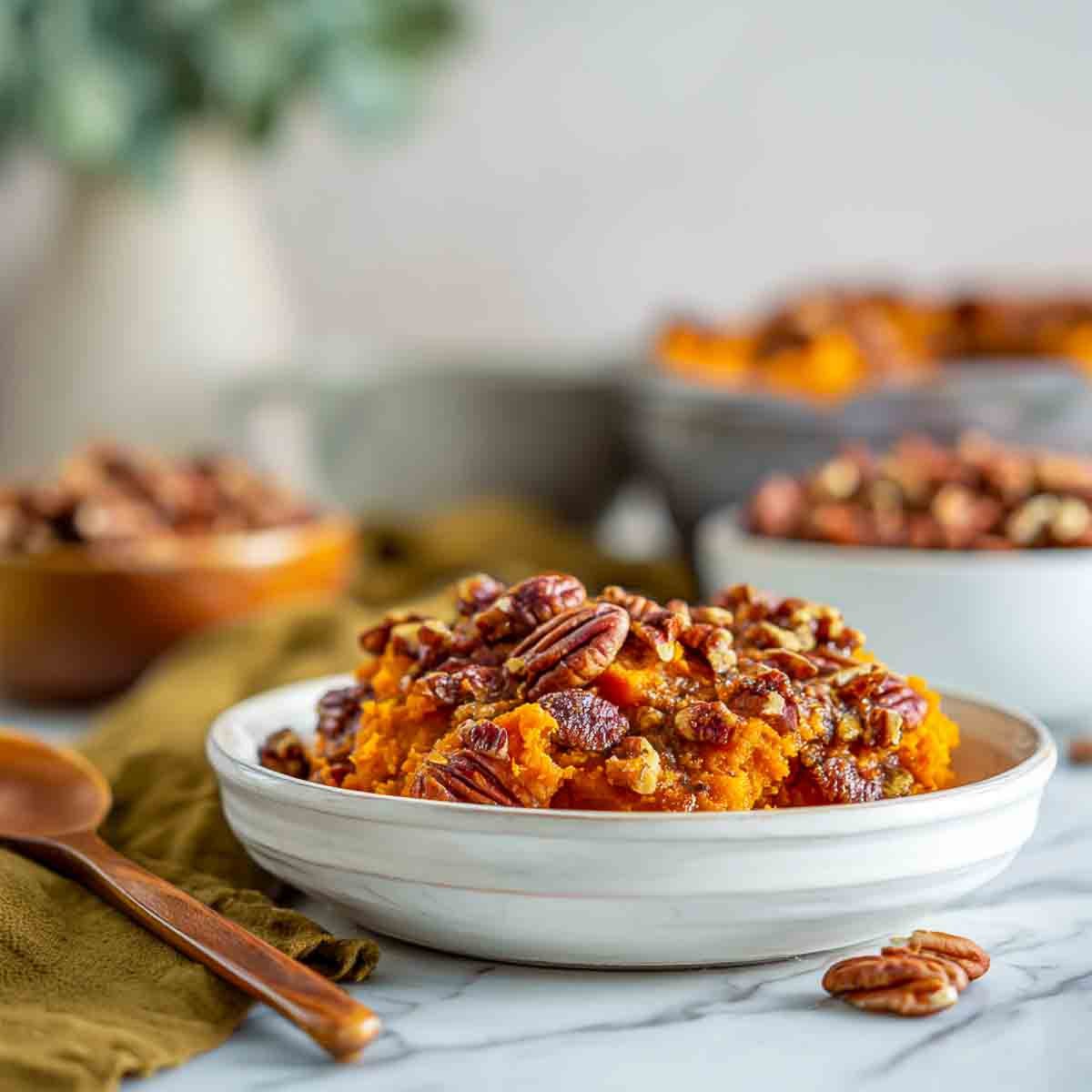 A plated serving of sweet potato casserole topped with glossy cinnamon pecans, with small bowls of pecans and a wooden spoon in the background.