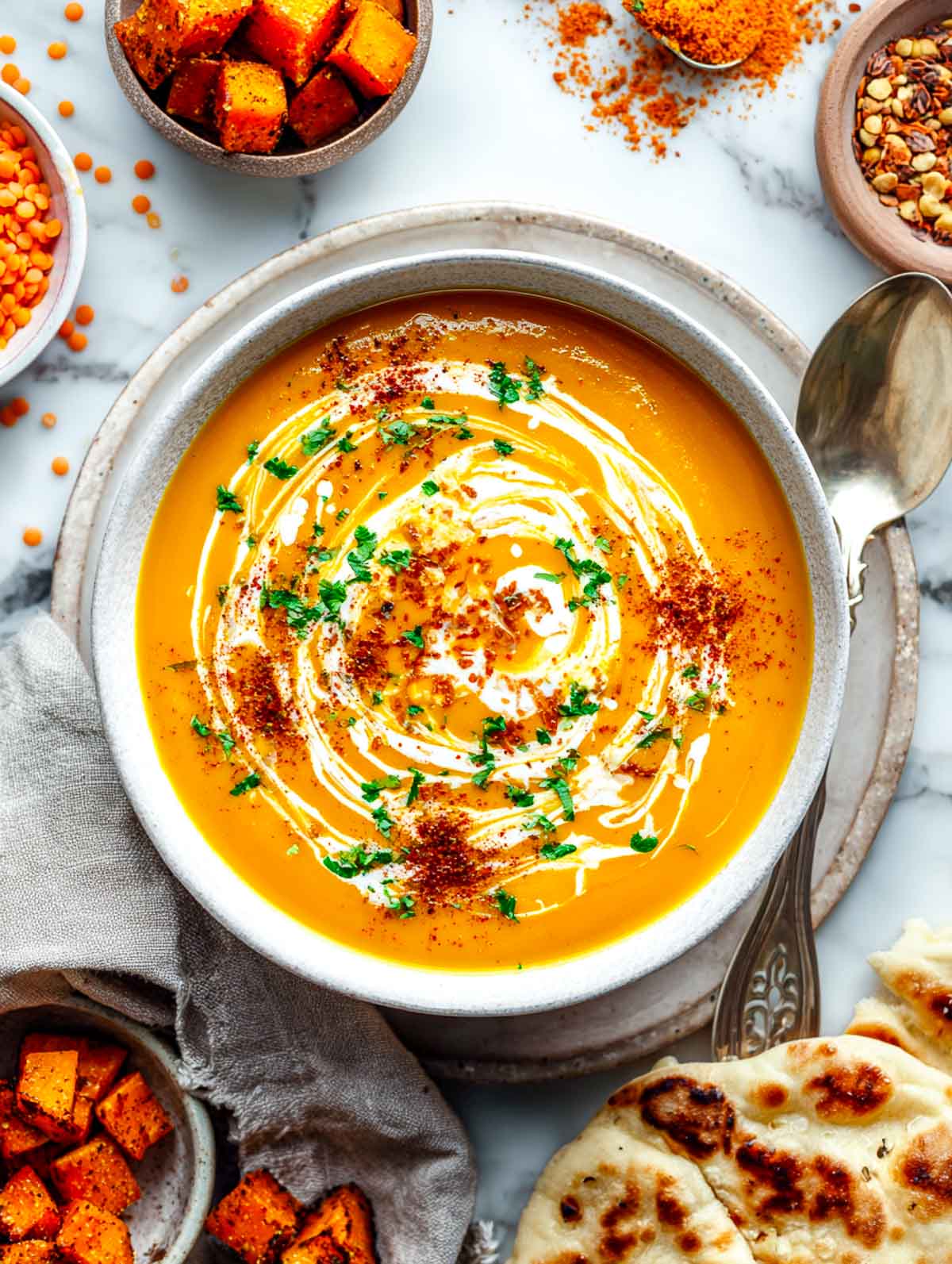 Curried sweet potato and butternut soup with coconut milk swirl, curry powder, and naan on a marble surface.