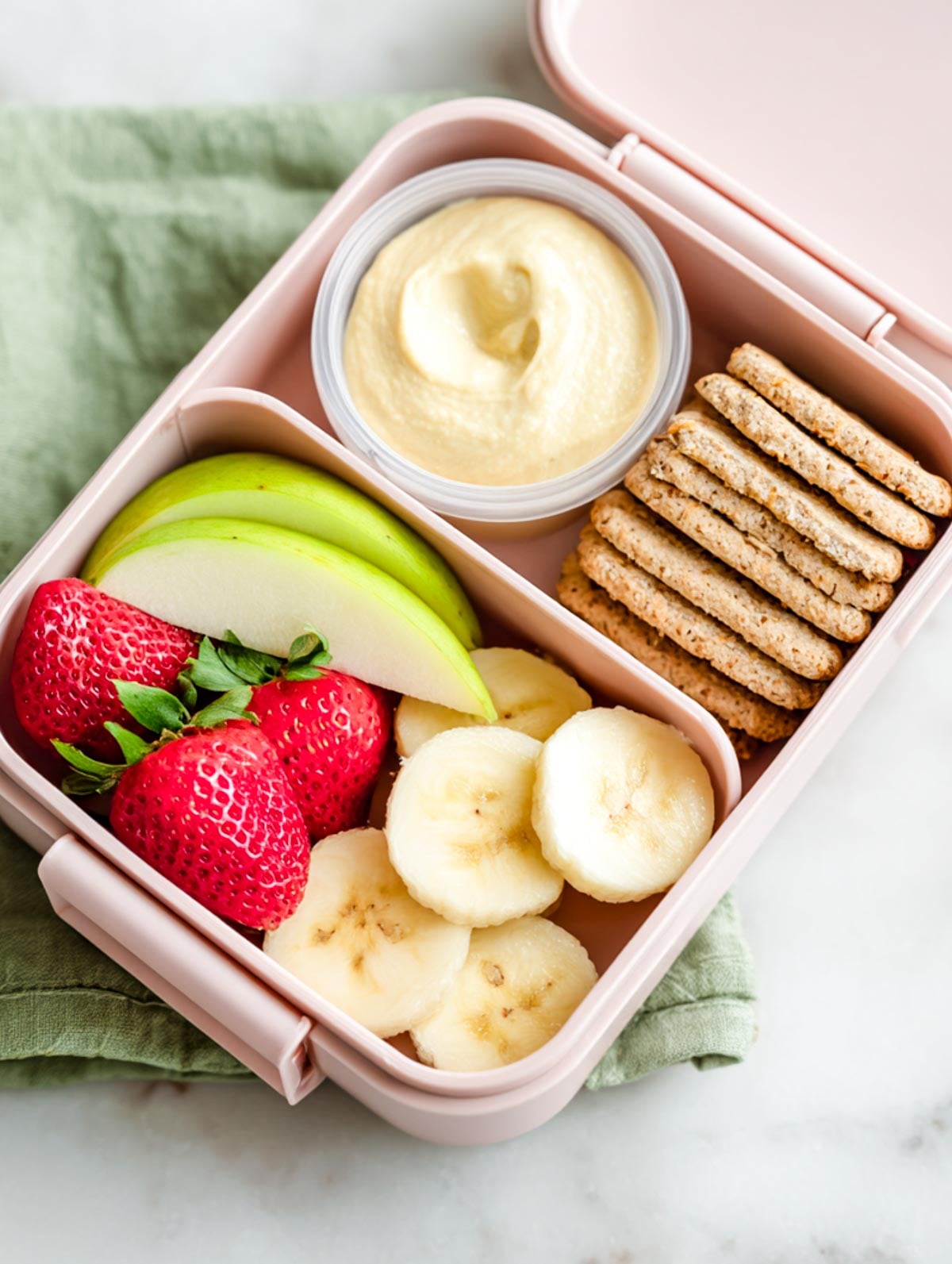 Fruit and hummus snack bento in a blush pink lunchbox with crackers, banana slices, apple wedges, and strawberries on a marble surface.