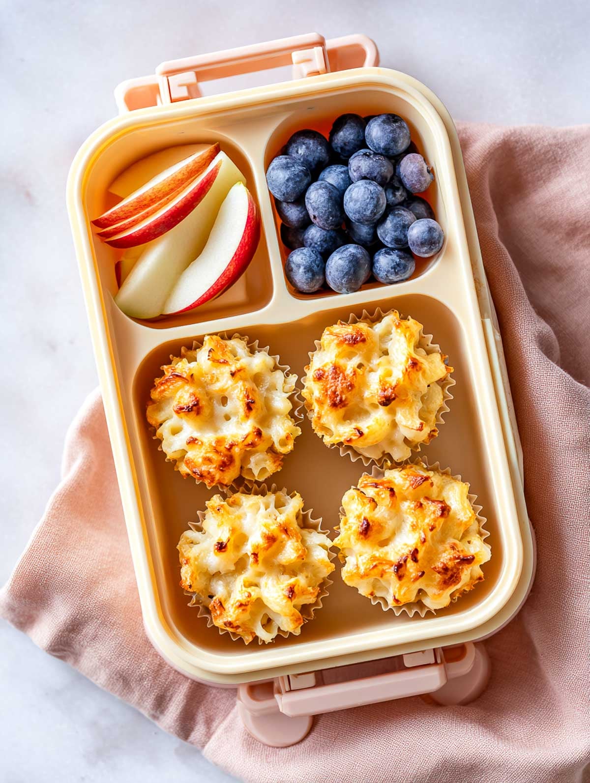 Hidden veggie mac and cheese bites in a pale yellow lunchbox with apple slices and blueberries on a marble surface.