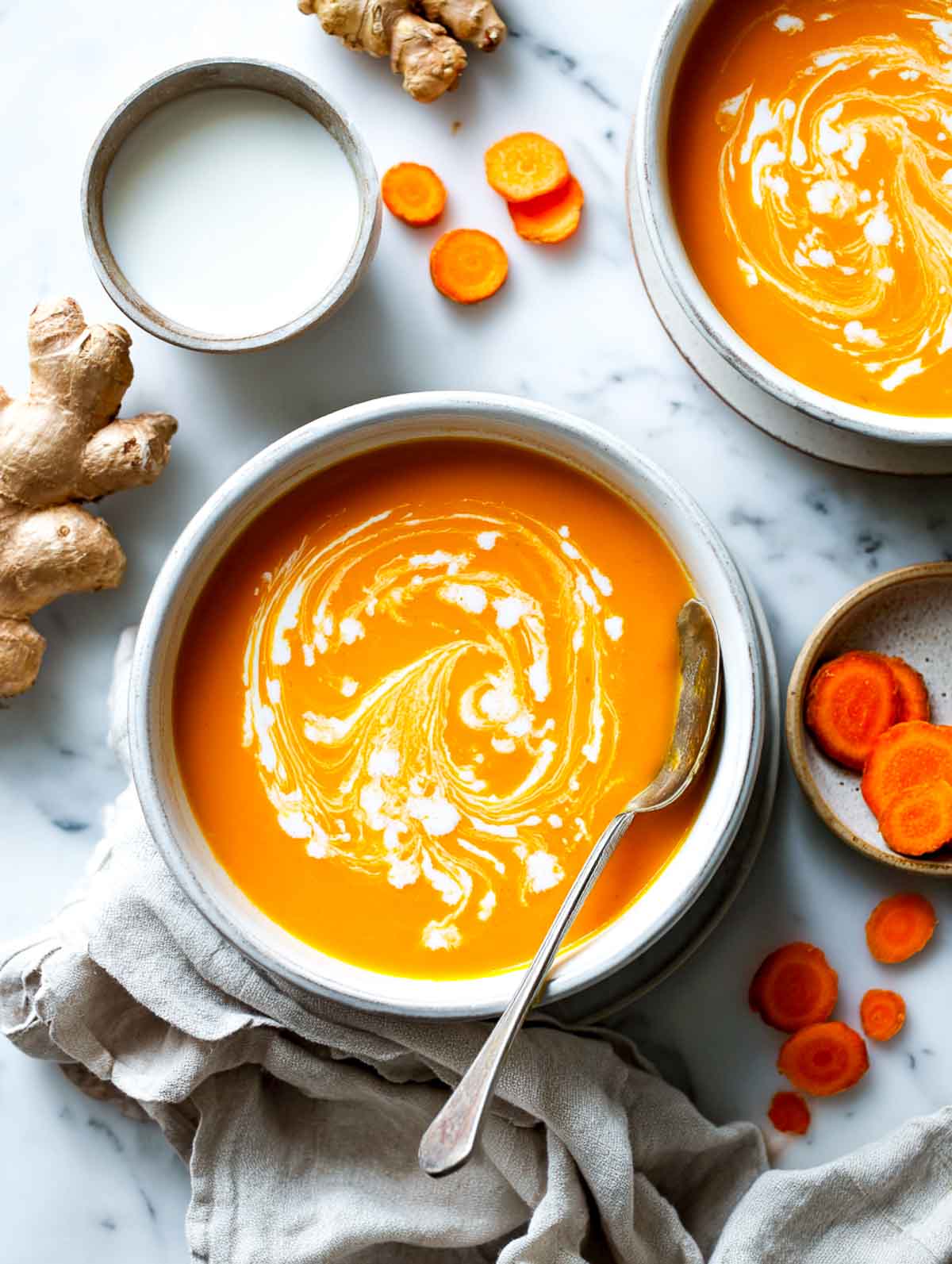 Vibrant orange sweet potato and carrot soup with a swirl of ginger coconut cream, served on marble with fresh ginger and carrots.