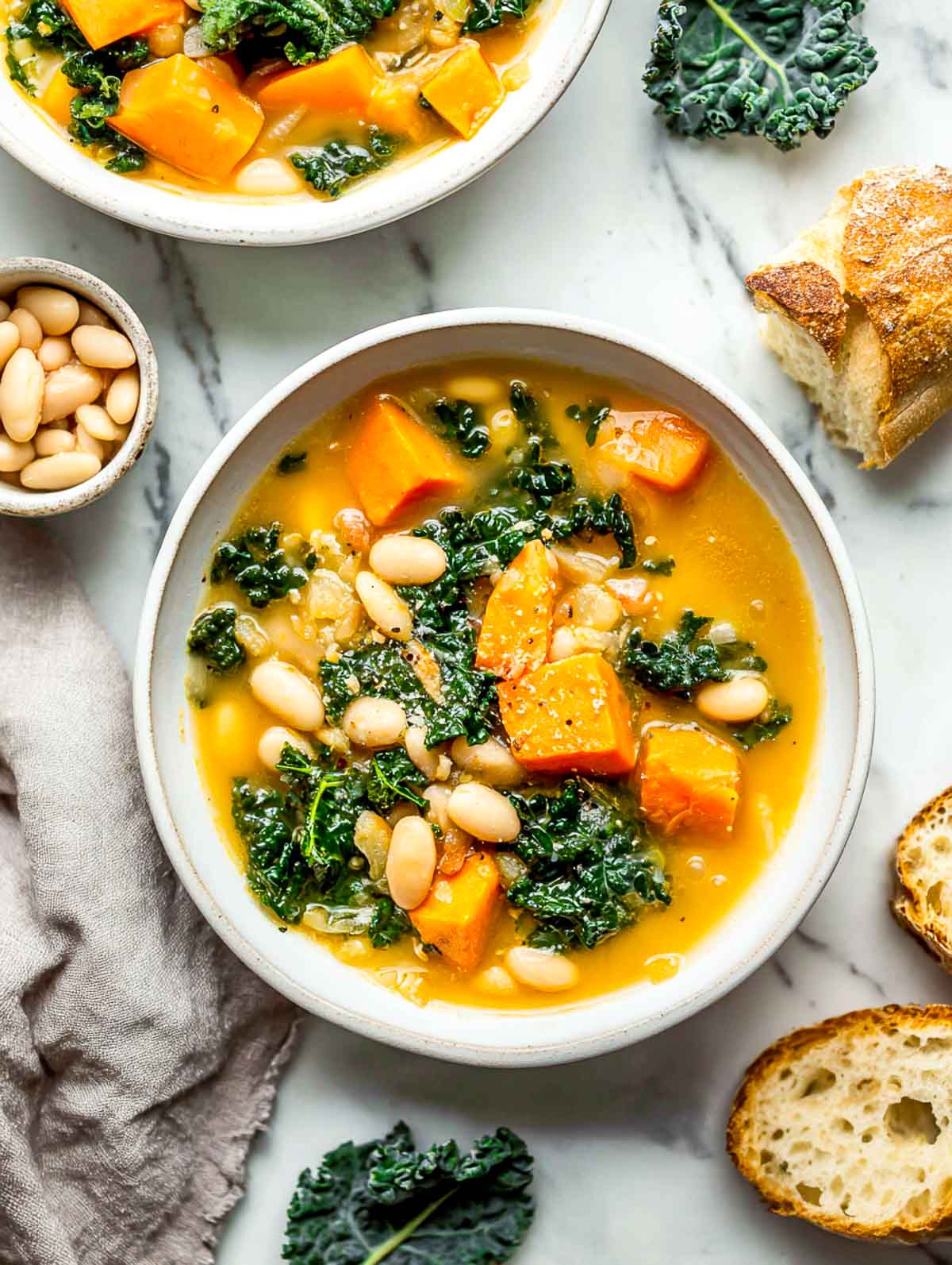 Hearty sweet potato and kale soup with white beans served in a white bowl on a marble surface with bread on the side.
