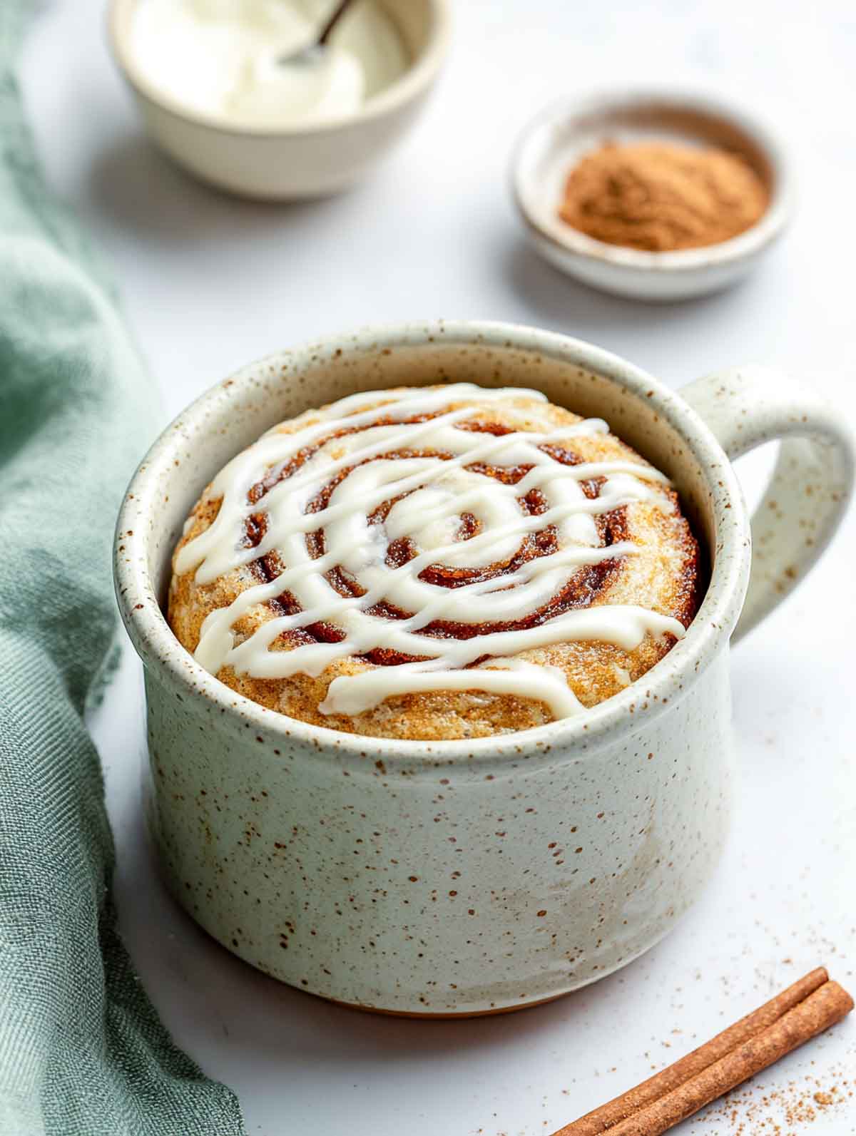 A warm keto cinnamon roll mug cake in a speckled mug with a neat cream cheese drizzle over a visible swirl.