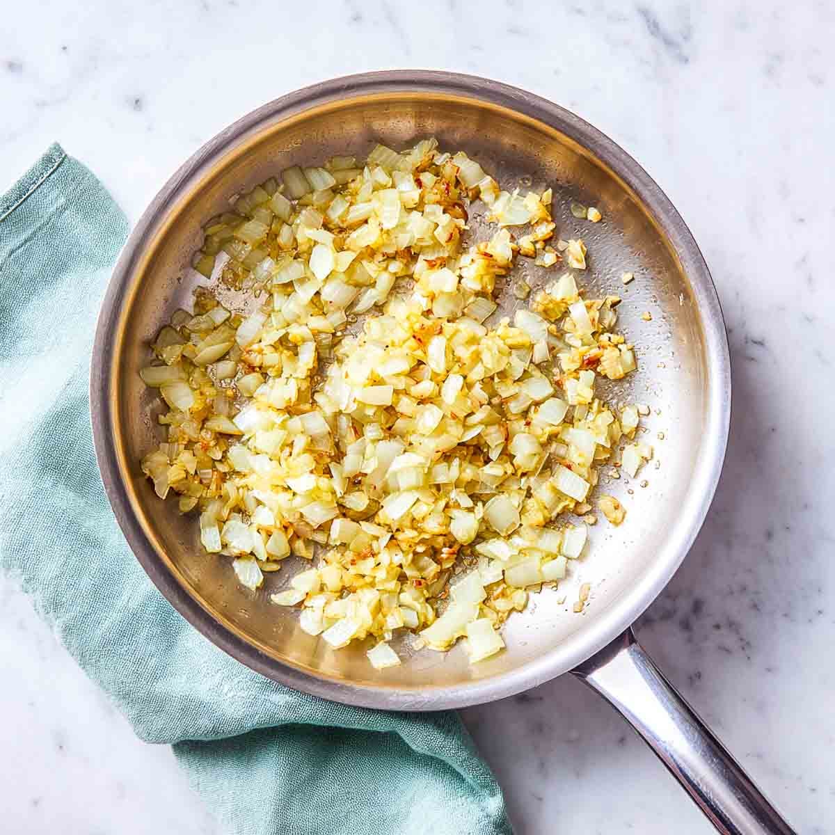 Onions, ginger, and garlic sautéing in a stainless steel pan on marble surface.