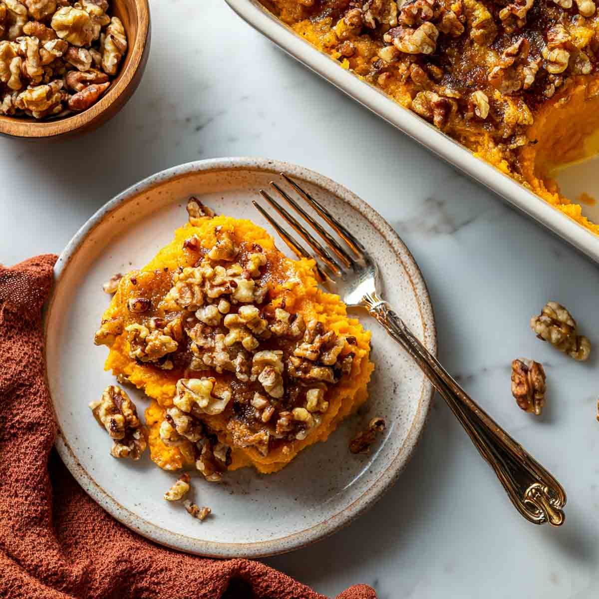 Top-down view of a plated serving of mashed sweet potato casserole topped with spiced walnuts, with the baking dish in the background on a marble surface.