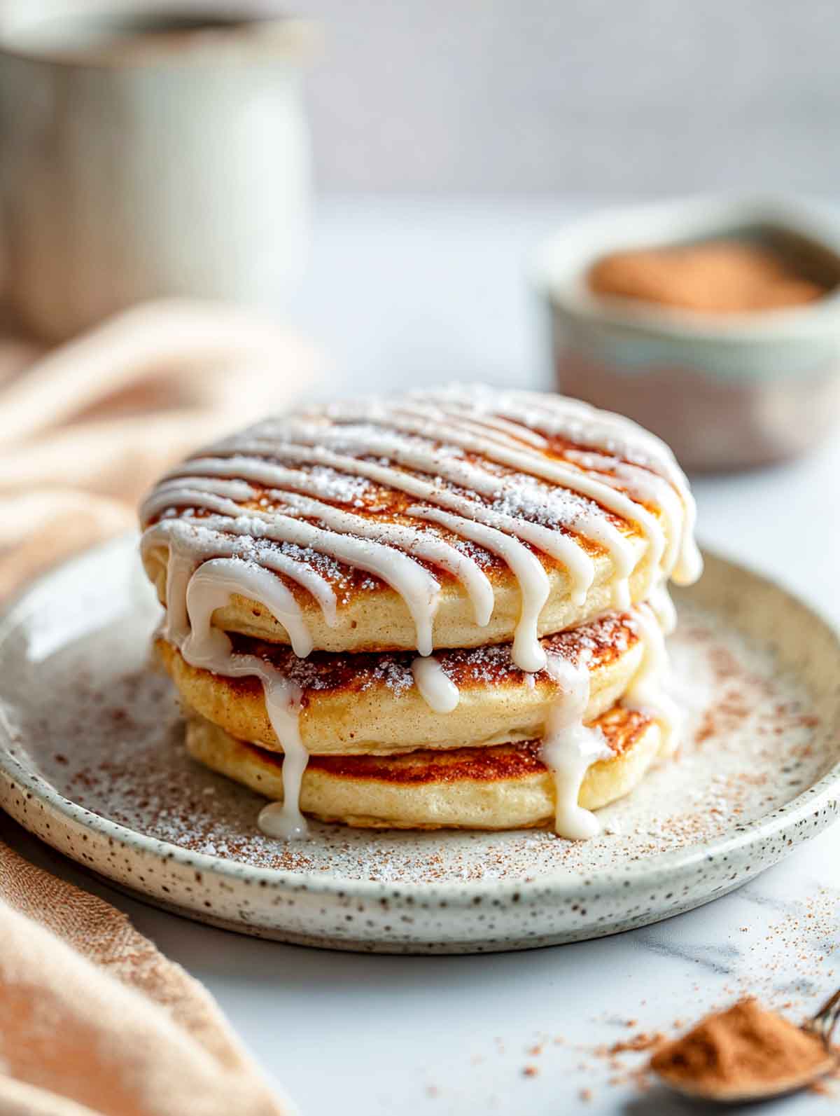 An angled view of a short stack of fluffy microwave pancakes drizzled with cinnamon glaze on a speckled plate.