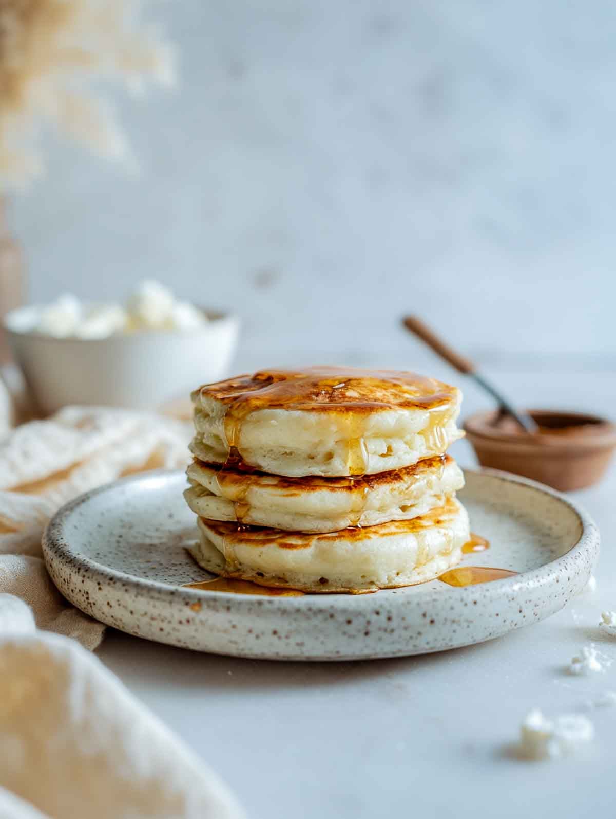 An angled view of a short stack of fluffy microwave cottage cheese pancakes drizzled with maple syrup on a speckled plate.