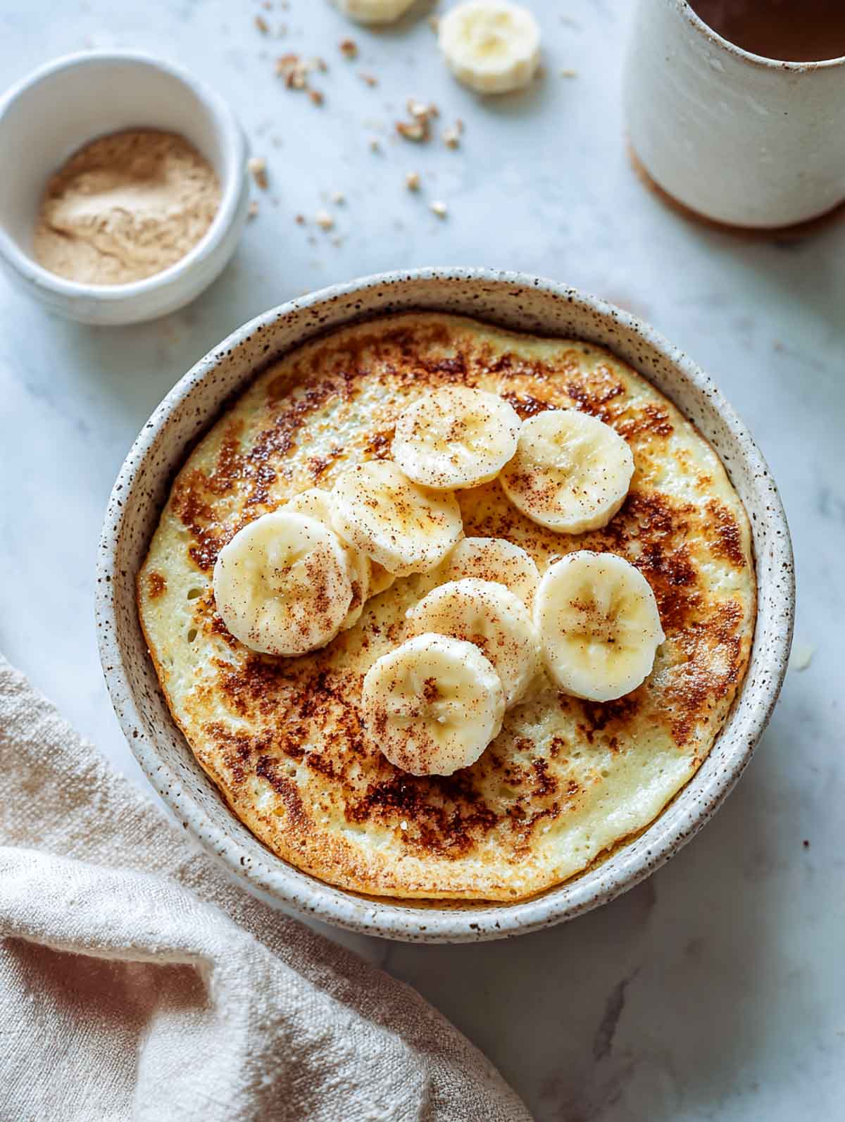 A top-down view of a fluffy microwave pancake served in a speckled bowl and topped with sliced bananas and cinnamon.