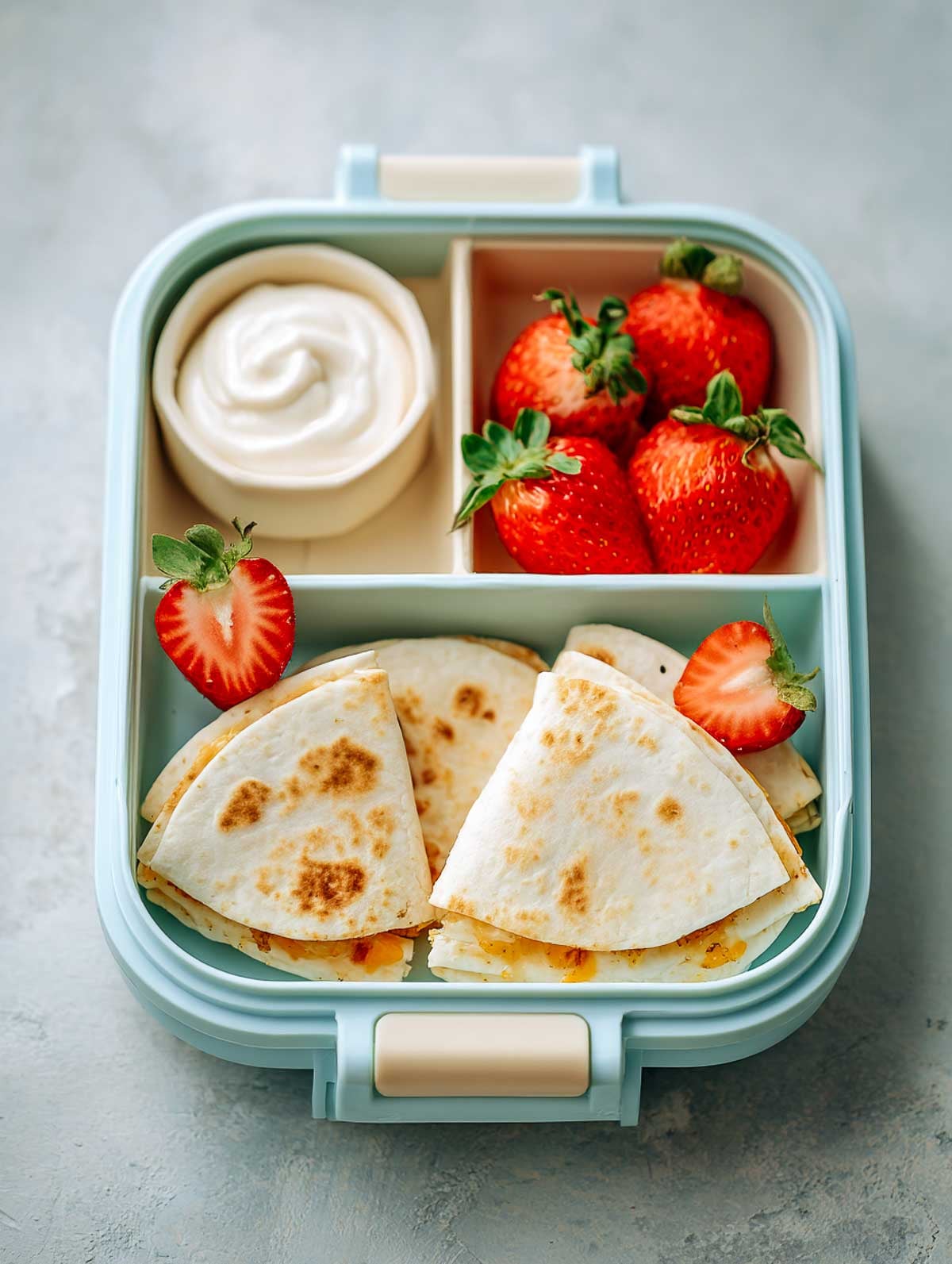 Mini veggie quesadilla bites packed in a mint-green lunchbox with strawberries and yogurt dip on a light surface.