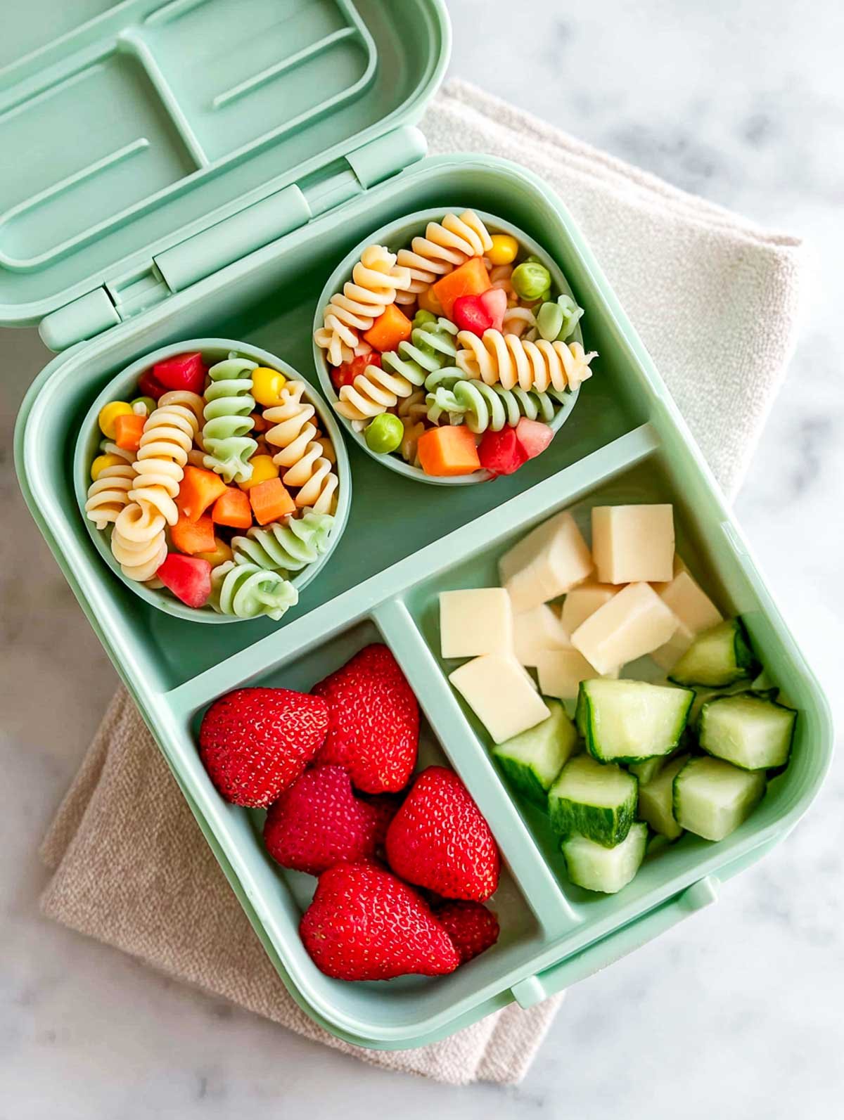 Rainbow pasta salad cups in a mint-green bento lunchbox with strawberries, cheese cubes, and cucumber pieces.