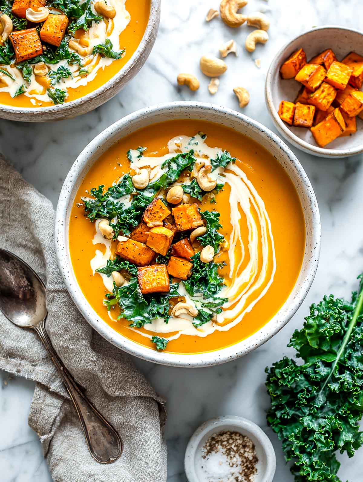 Roasted sweet potato and kale soup topped with garlic cashew cream, roasted cubes, and whole cashews on a marble surface.