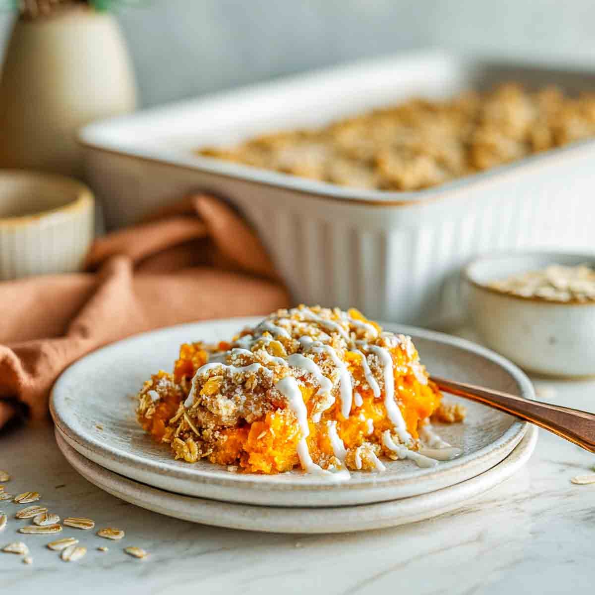 A plated serving of sweet potato casserole topped with oat crumble and yogurt drizzle, with the baked dish in the background on a marble counter.