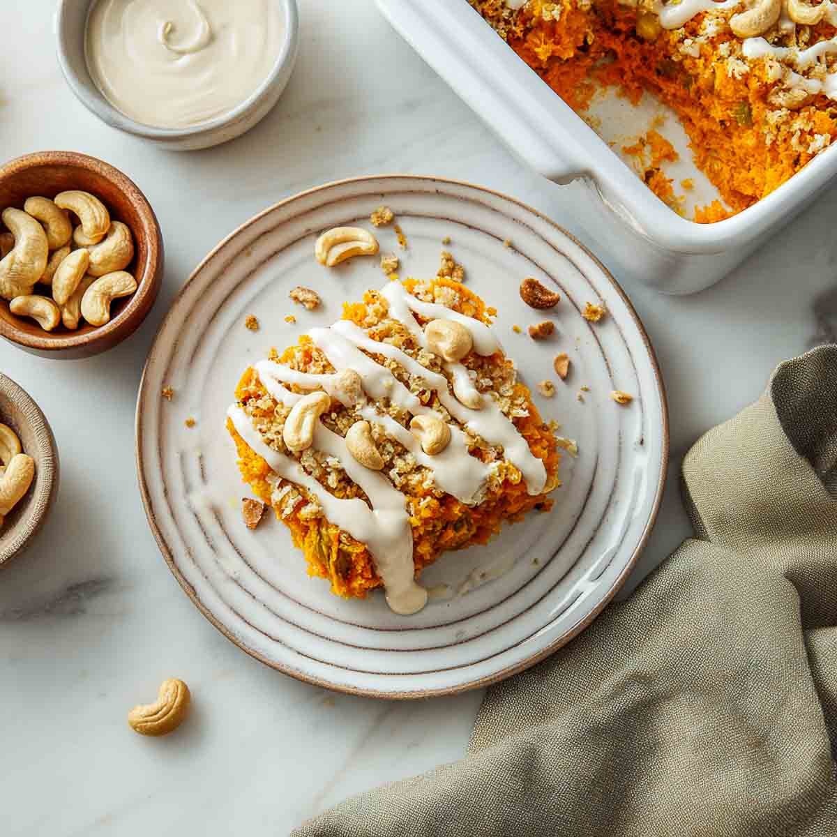 Top-down view of a plated serving of sweet potato casserole drizzled with spiced cashew cream sauce, with the baking dish in the background and scattered cashews on a marble countertop.