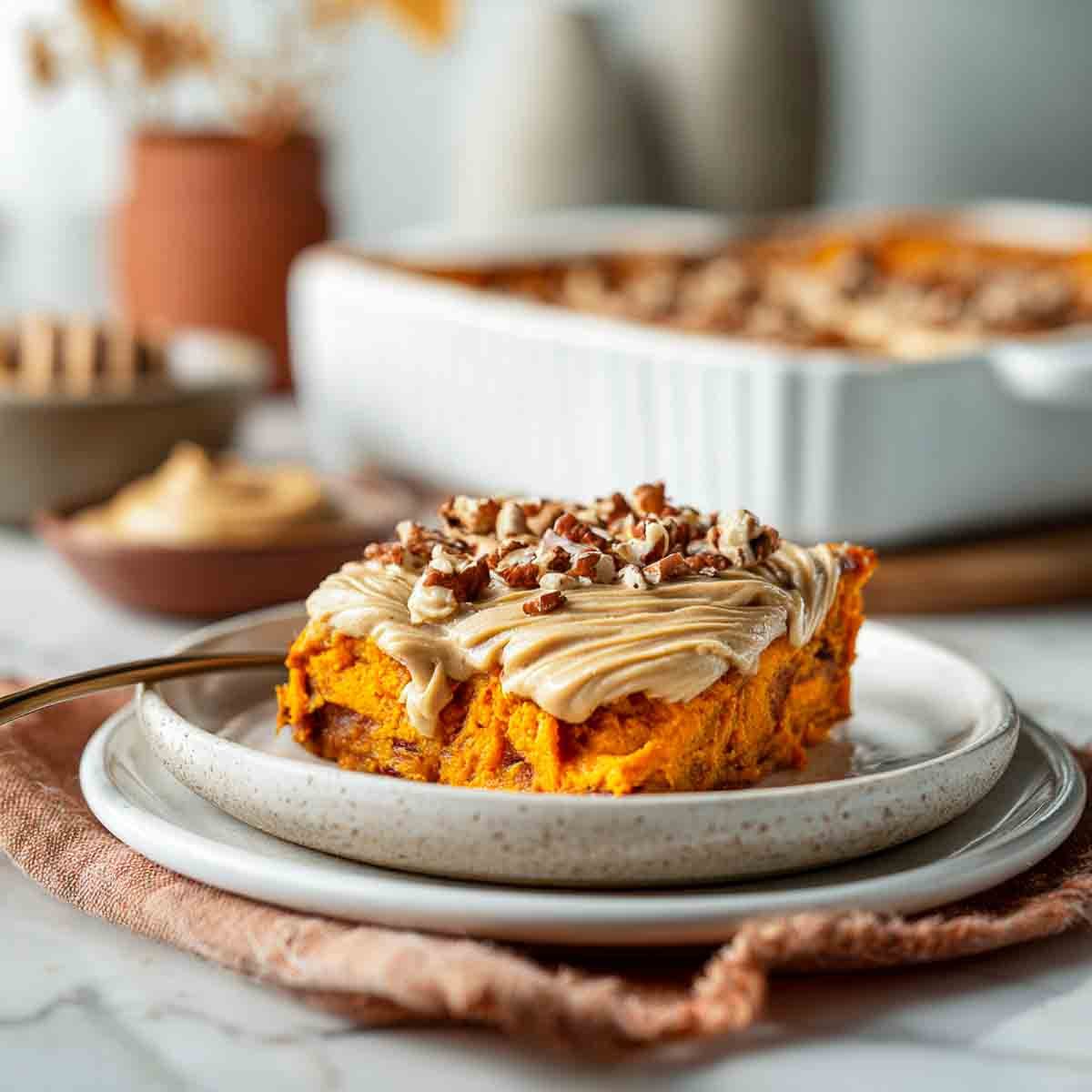 A plated serving of vegan sweet potato casserole topped with almond butter swirl and chopped pecans, with the baking dish in the background.