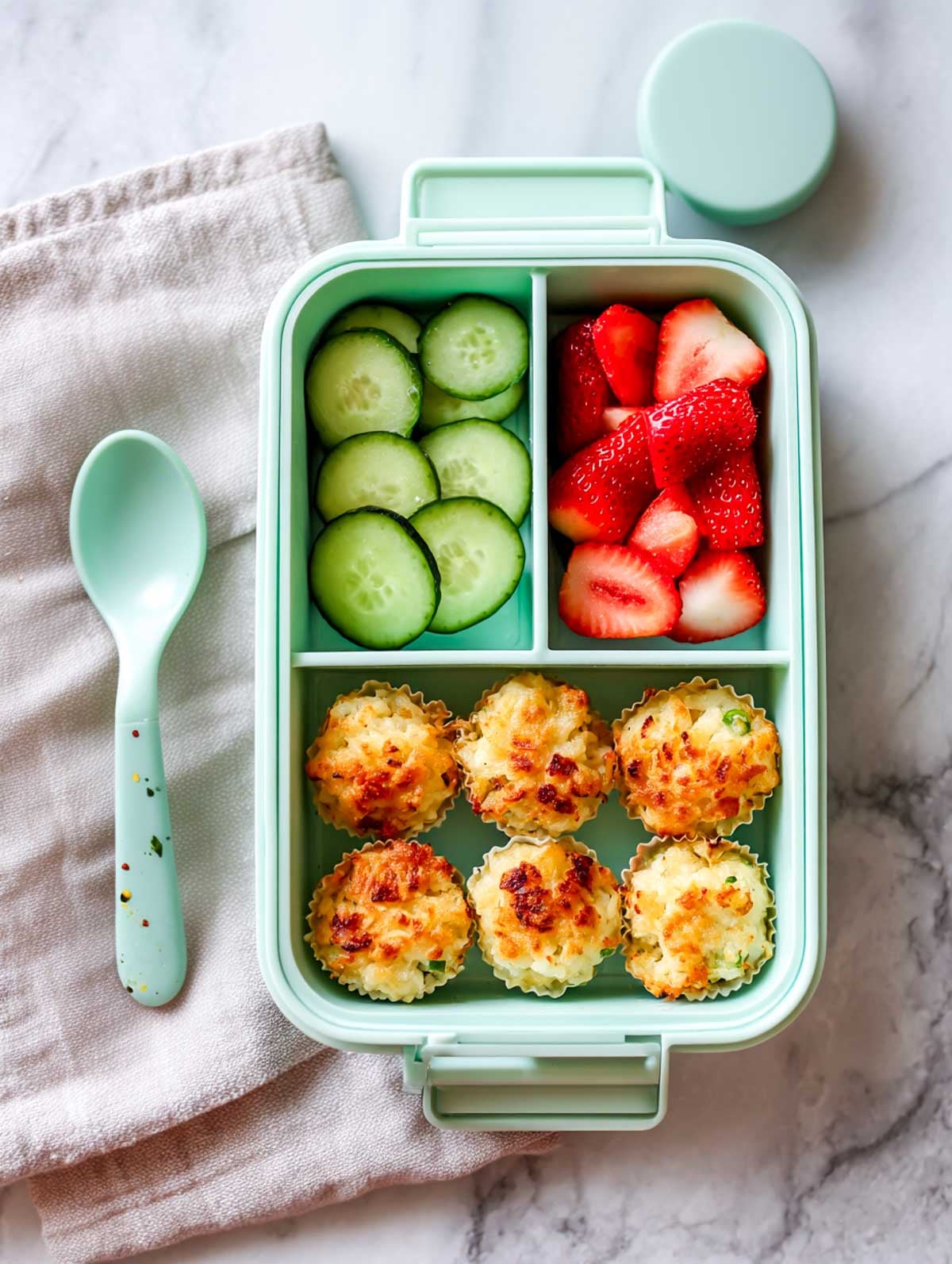 Veggie tater tot cups packed in a mint-green lunchbox with cucumber slices and strawberries on a marble surface.