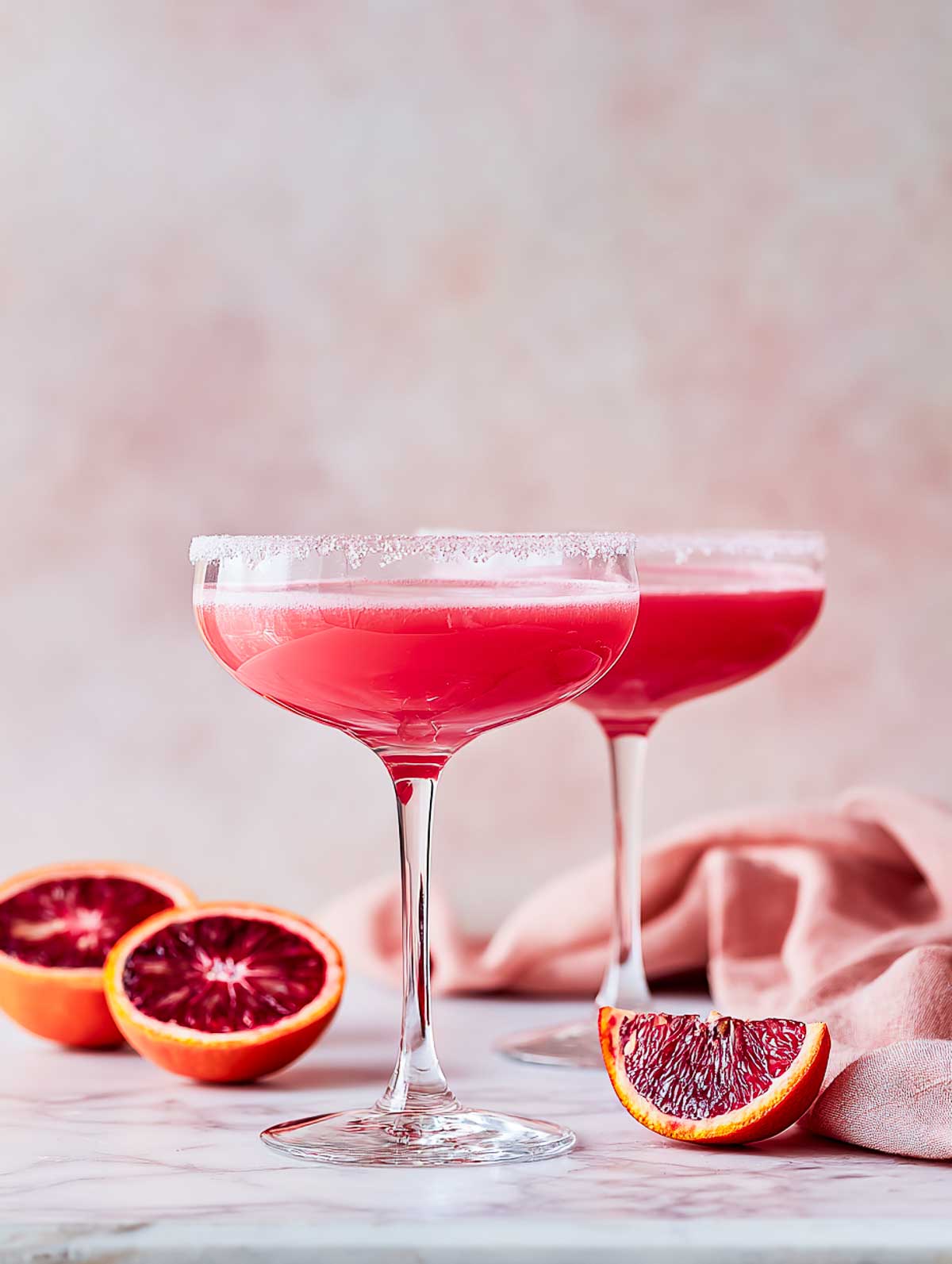 Side view of two blush pomegranate margaritas in coupe glasses on a white marble surface.