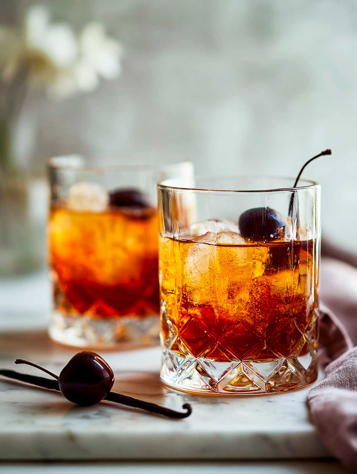 Side view of two cherry vanilla old fashioneds in rocks glasses on a white marble surface.