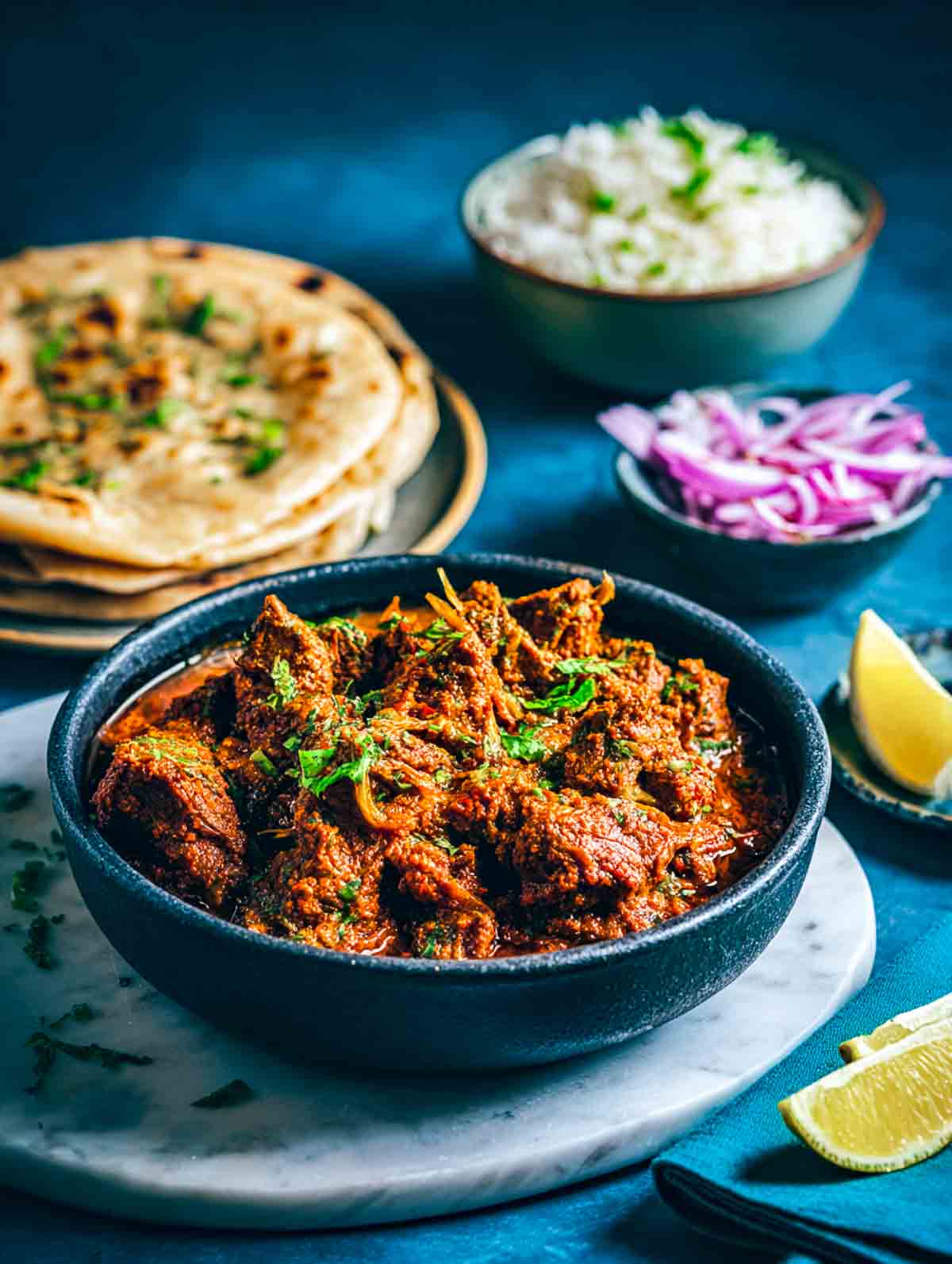 A bowl of lamb bhuna curry served with roti, rice, onions, and lemon wedges.