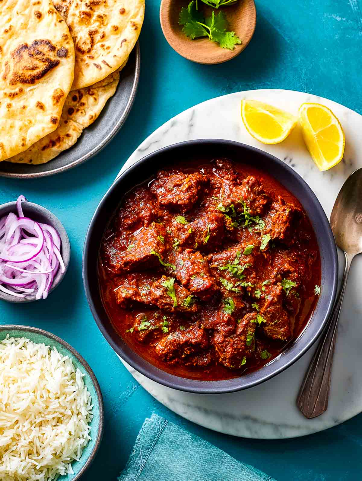 Lamb bhuna curry in a black bowl on marble, garnished and surrounded by sides.