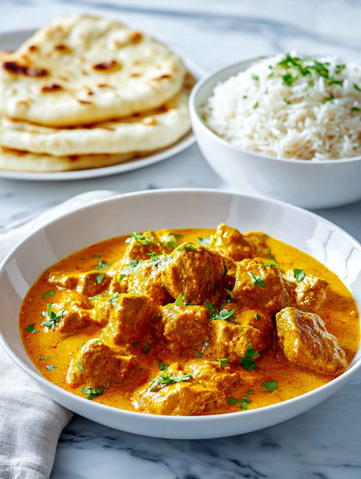 Side view of lamb korma curry served in a white bowl with rice and naan in the background.